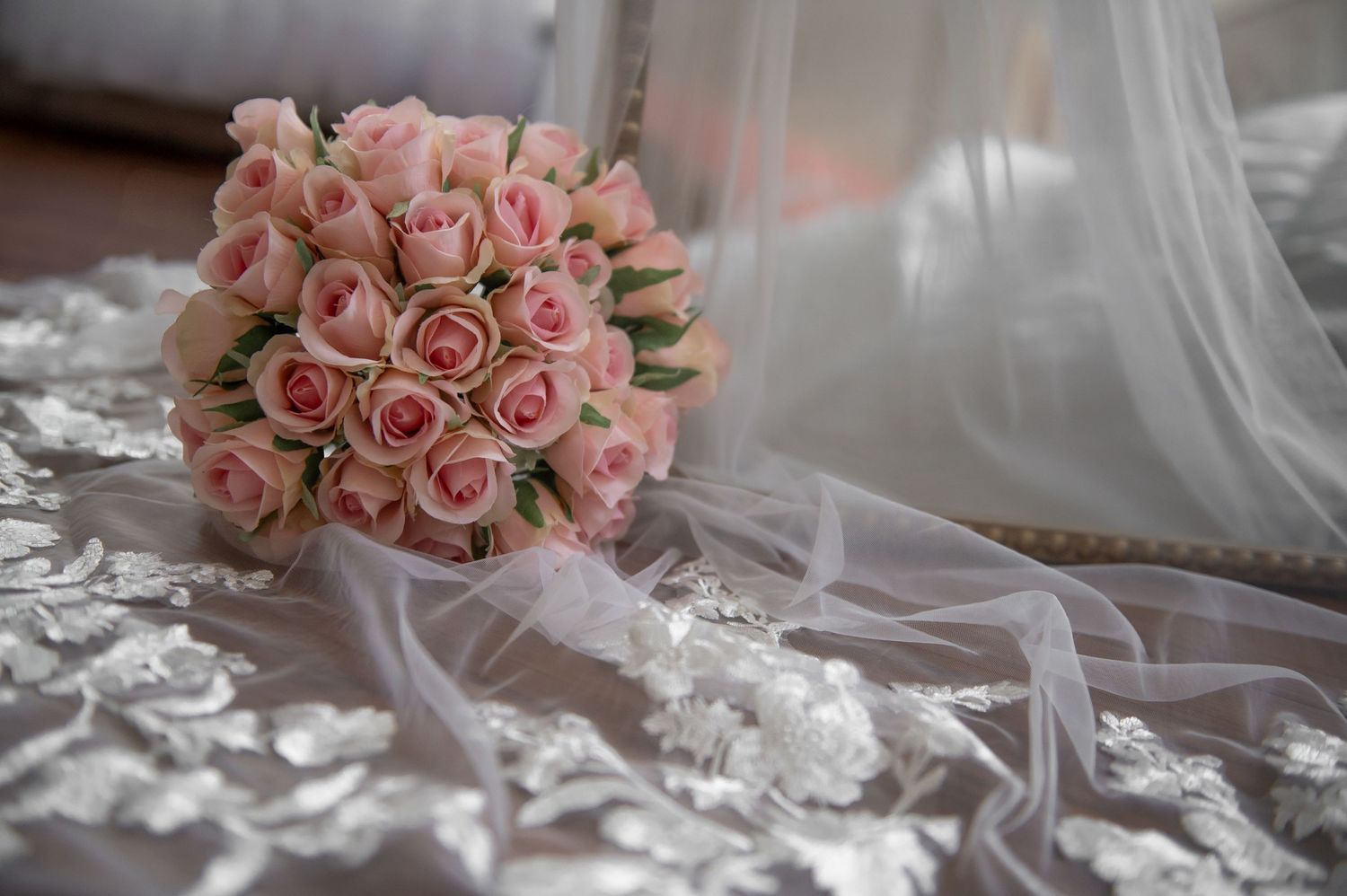 A delicate pink rose wedding bouquet rests on white lace with flowing tulle veil in soft natural lighting.