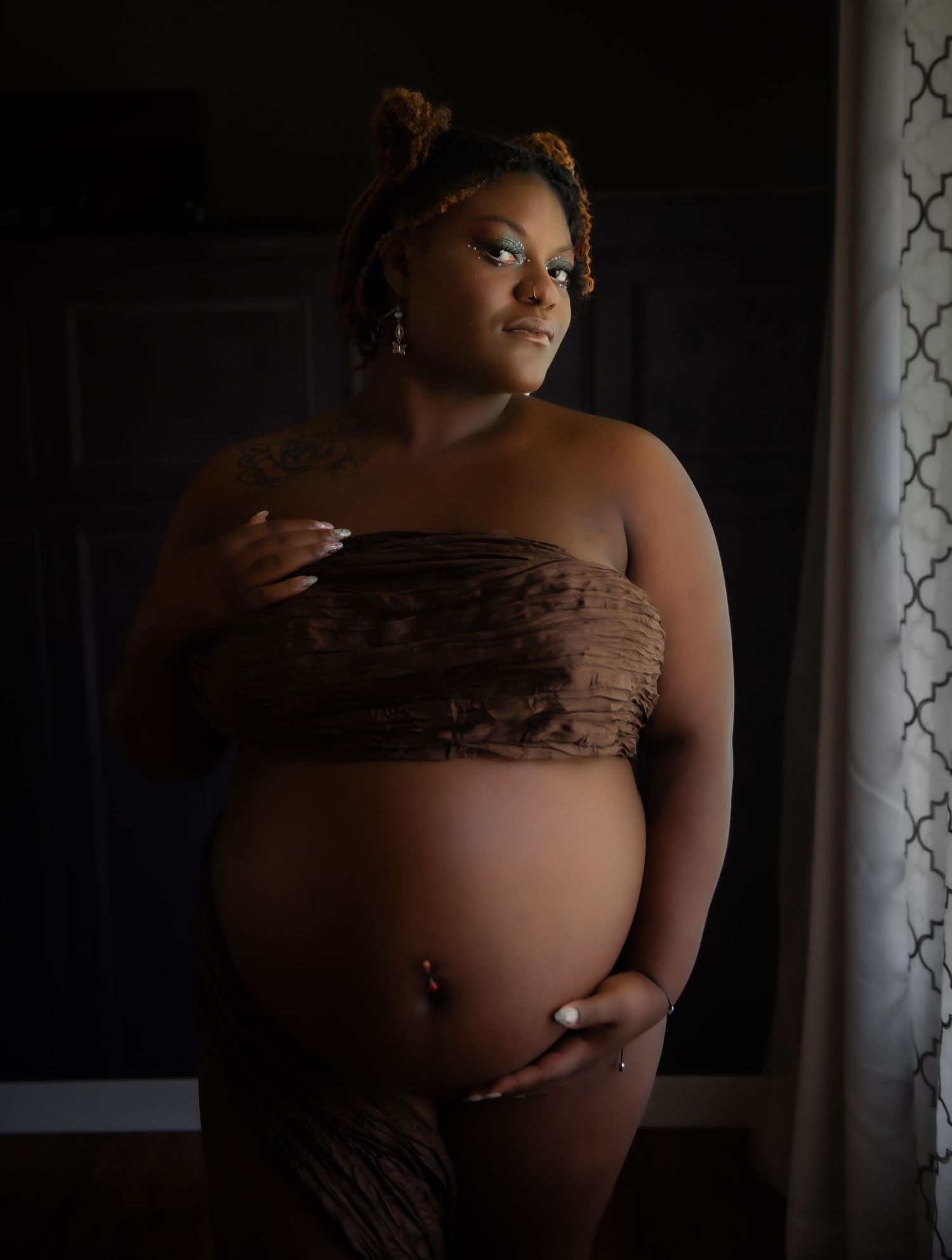 Artistic maternity photo showing a woman in brown strapless attire posing against a dark background near curtains.