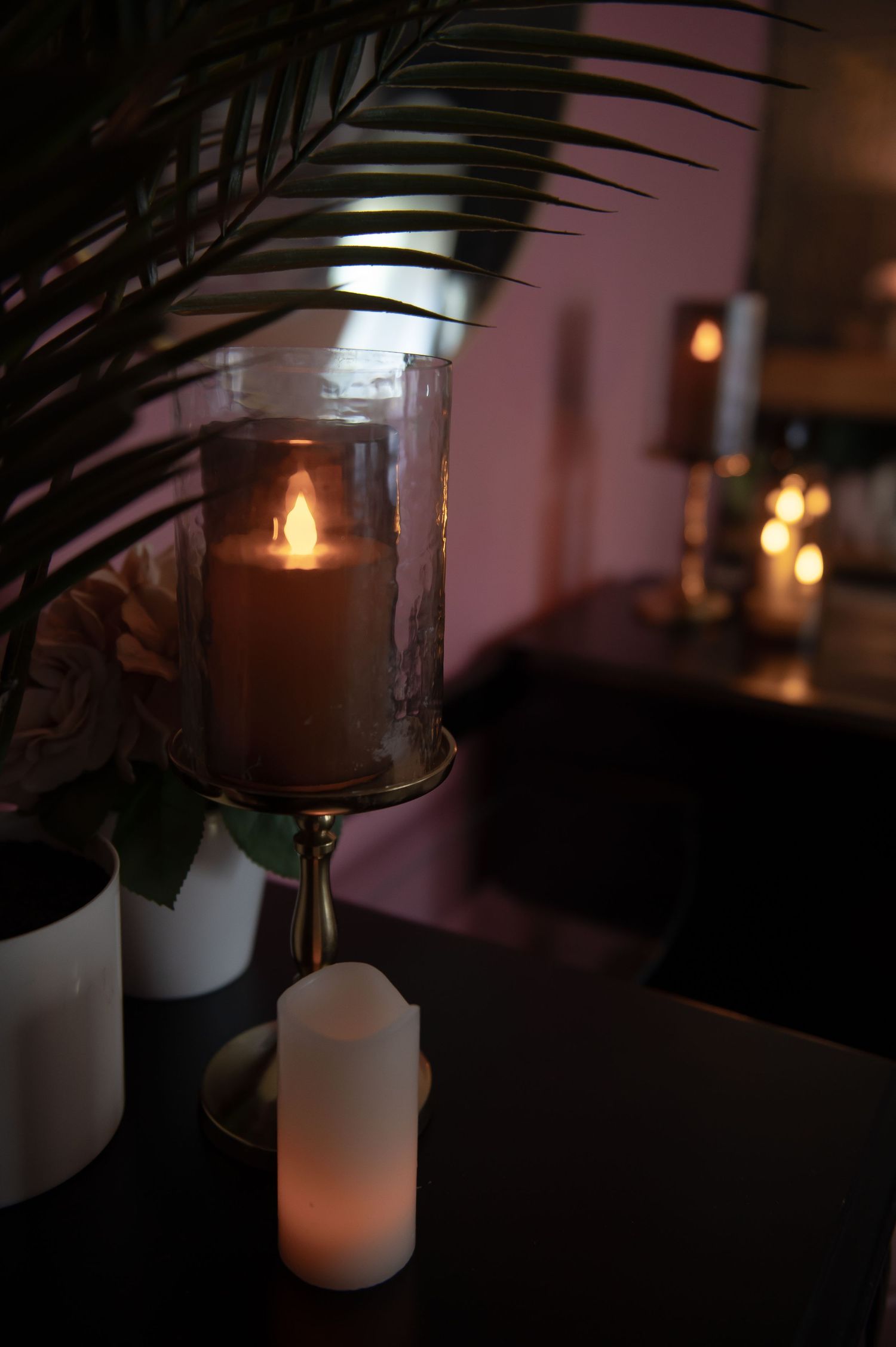 A lit candle glows warmly in a glass holder with palm fronds and white pillar candles creating a cozy boudoir set ambiance.