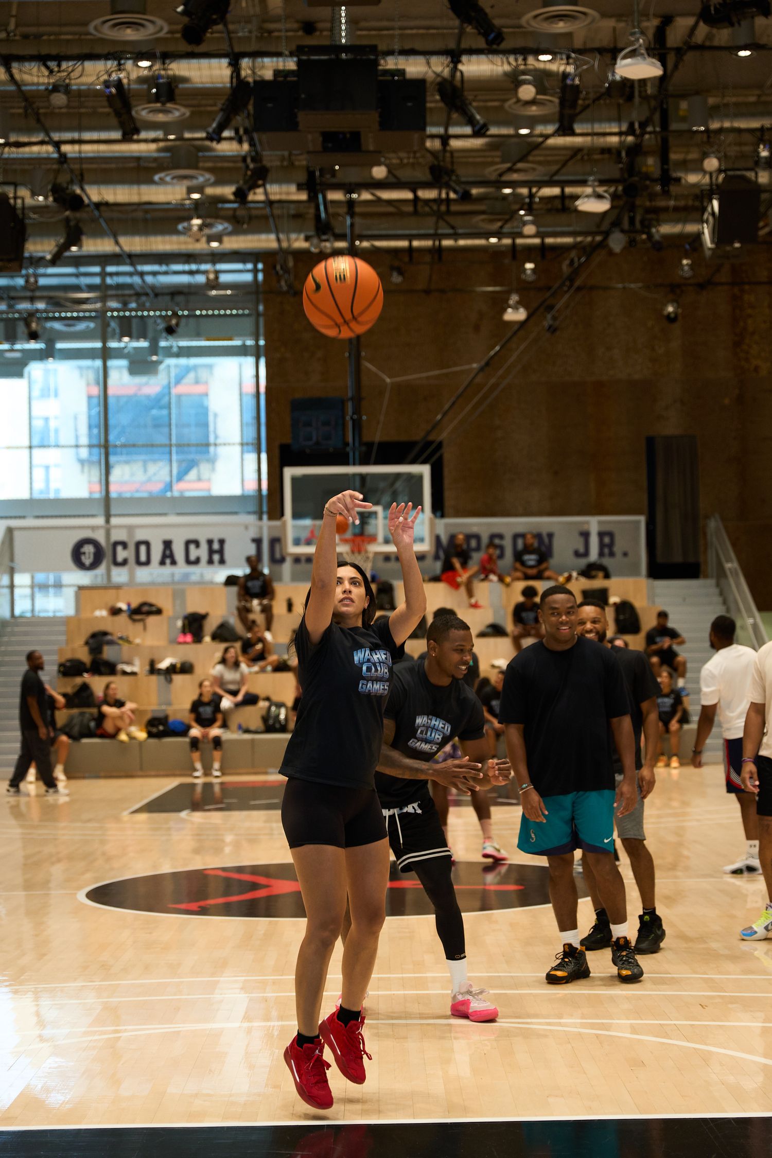 A basketball game in progress on an indoor court with players practicing and a basketball visible in the air.