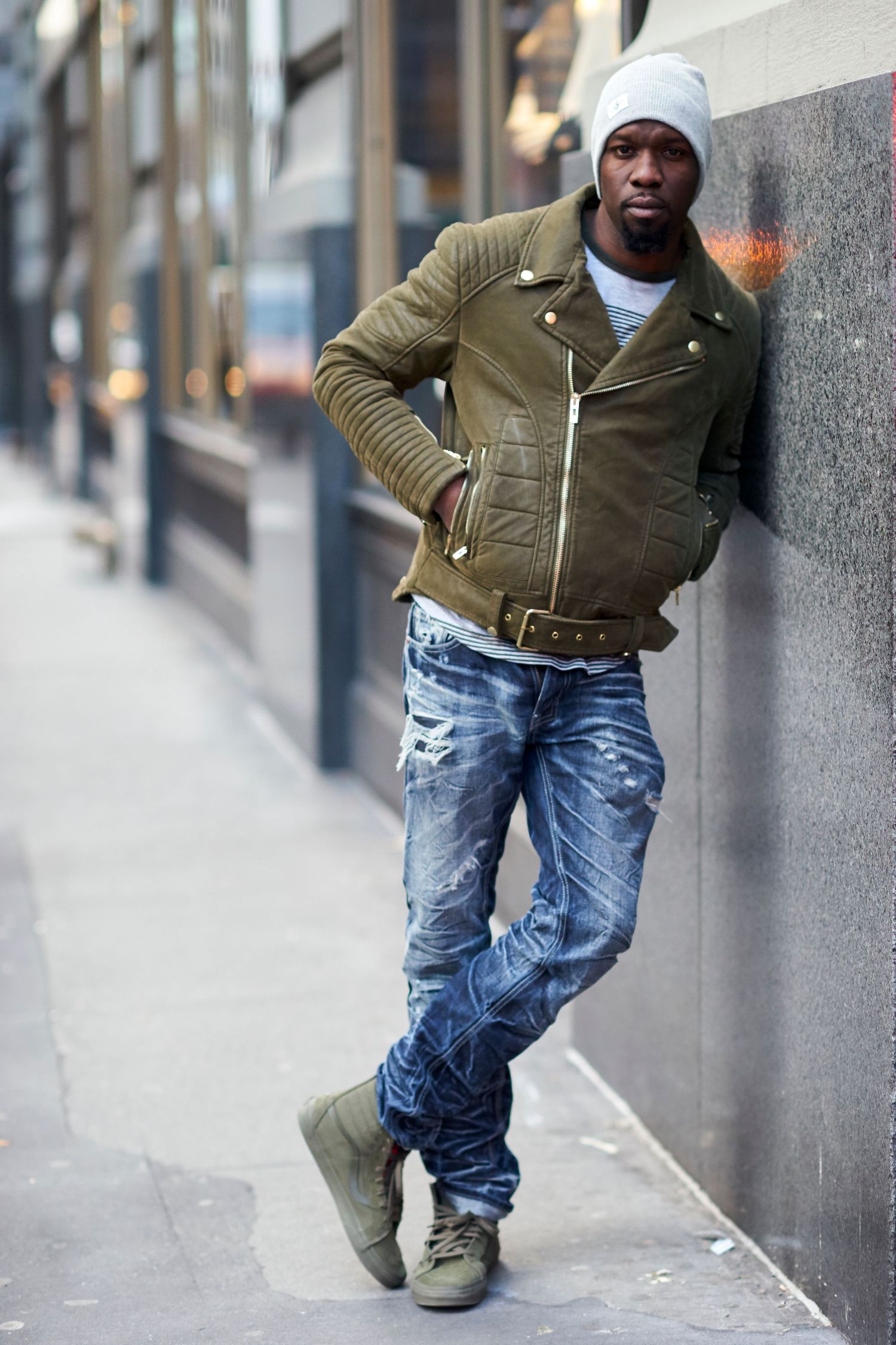 Street style fashion with olive green moto jacket, distressed denim jeans, and matching boots against urban wall.