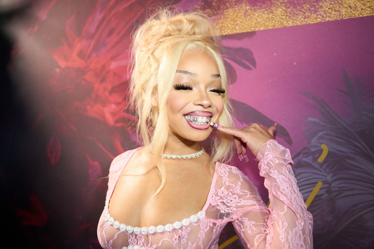 Performer wearing a pink feathered gown and blonde hair performing on stage with dramatic red lighting effects.