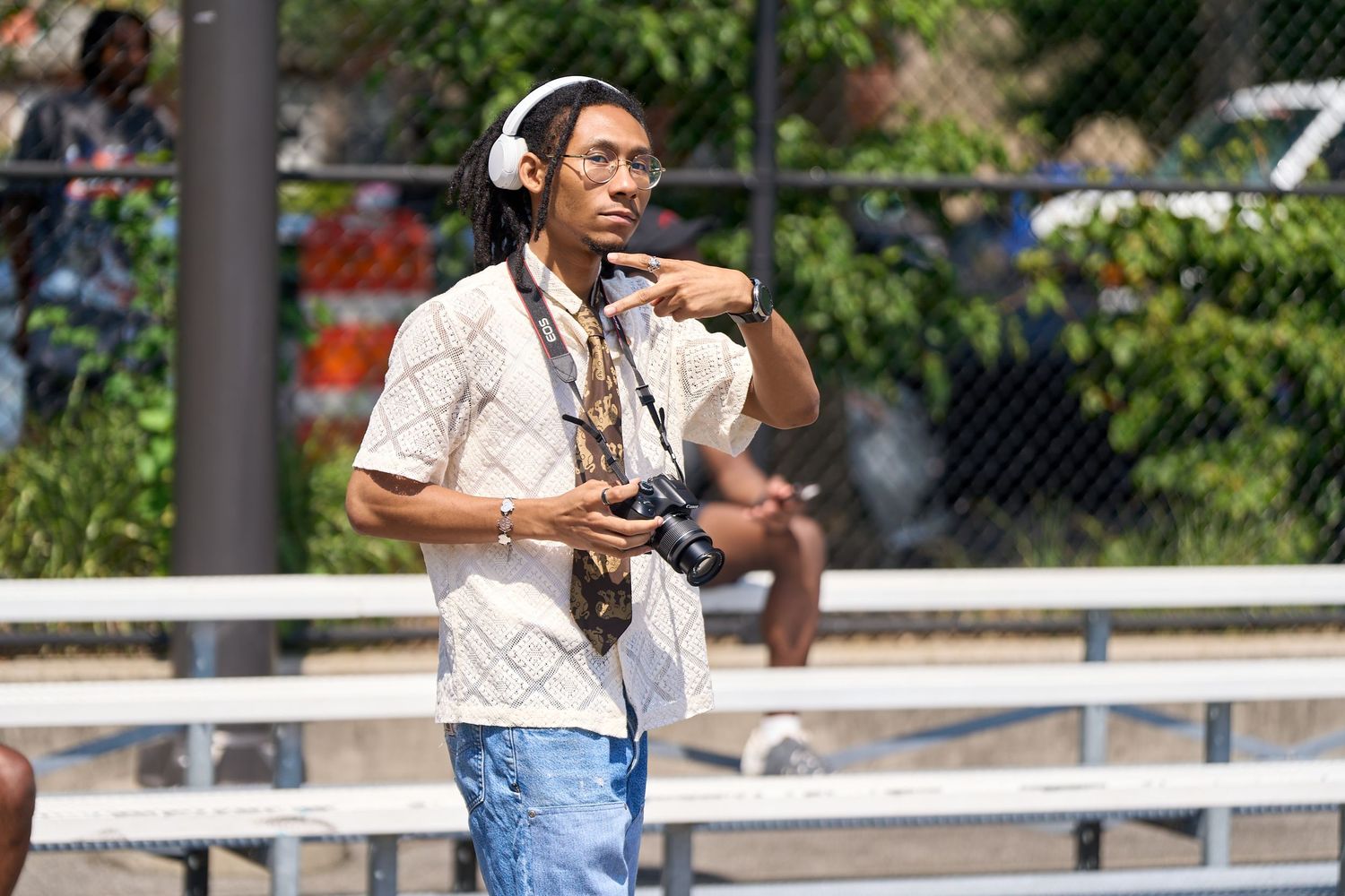 Photographer wearing a light patterned shirt and headphones stands on bleachers holding camera equipment.