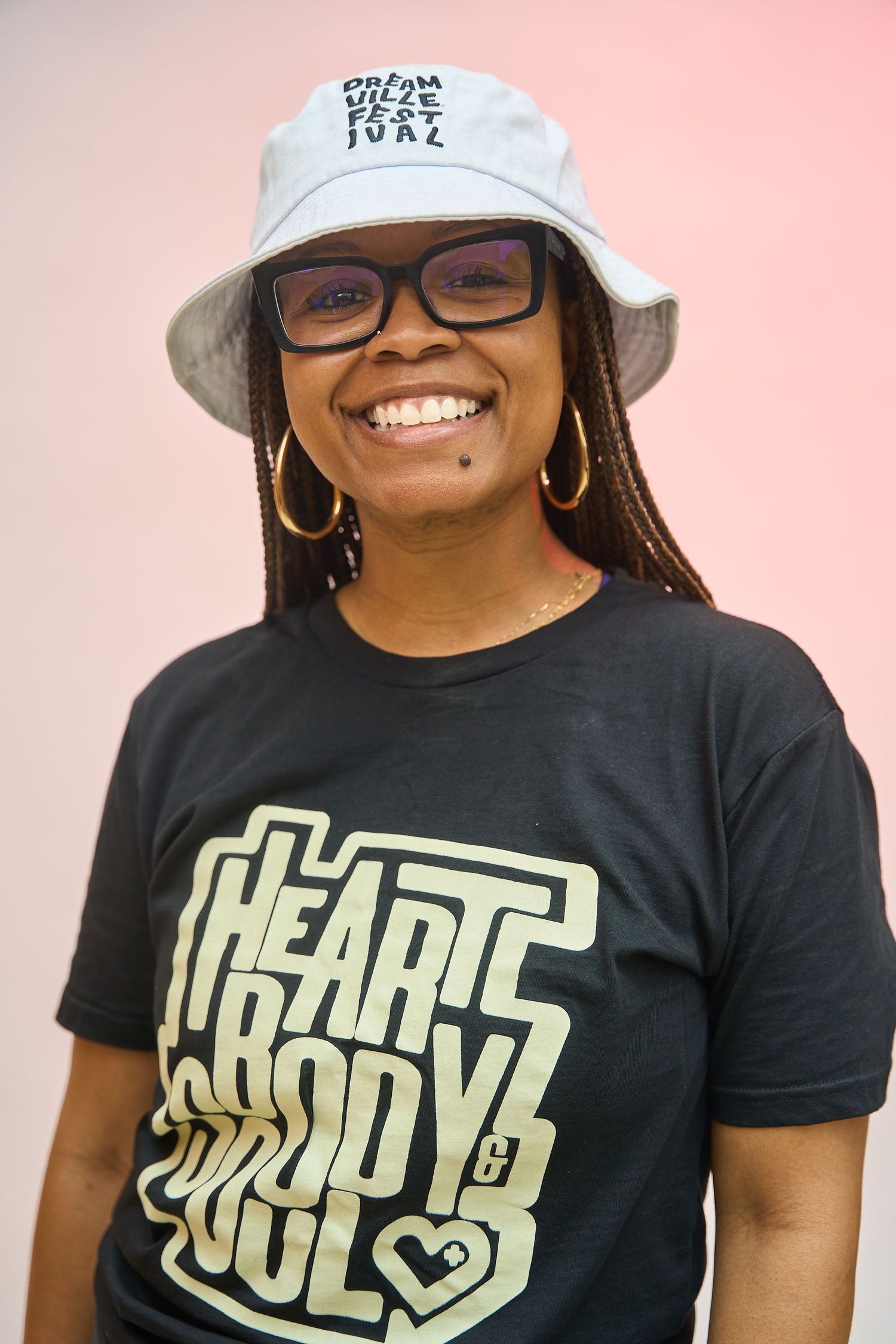 Smiling woman wearing glasses, bucket hat, and Heart Body Soul black t-shirt against pink background.
