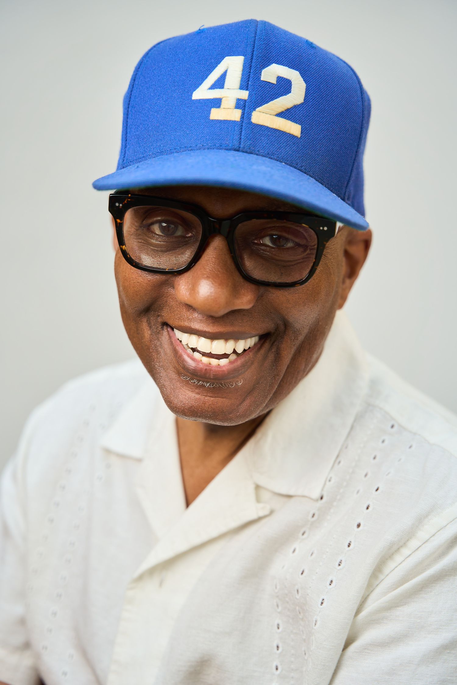 Smiling man wearing blue baseball cap with number 42 and black glasses, dressed in white shirt.