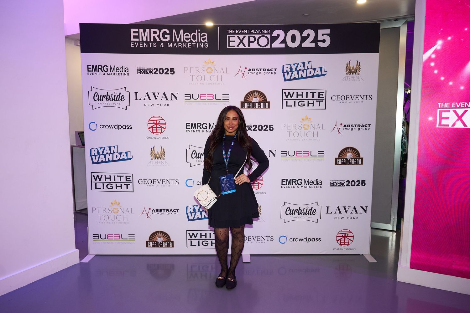 Person in black outfit poses against sponsor wall backdrop with corporate logos at promotional event.