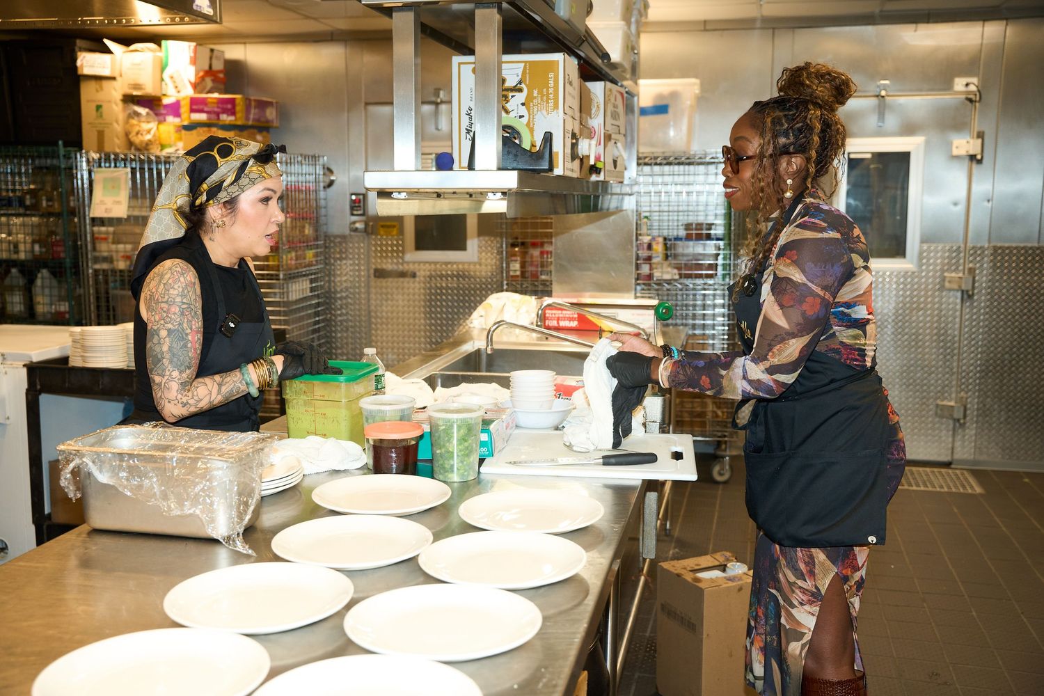 Two women talk across a stainless steel counter in a commercial kitchen, with white plates, food containers, and shelves in the background.