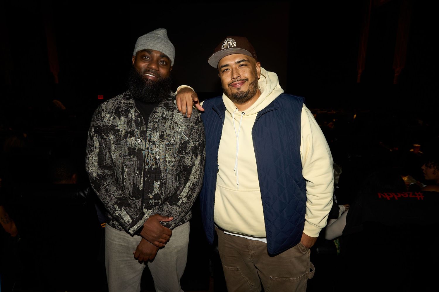 Two men pose together smiling at an indoor event, one wearing a beanie and patterned shirt, the other in a navy hoodie.