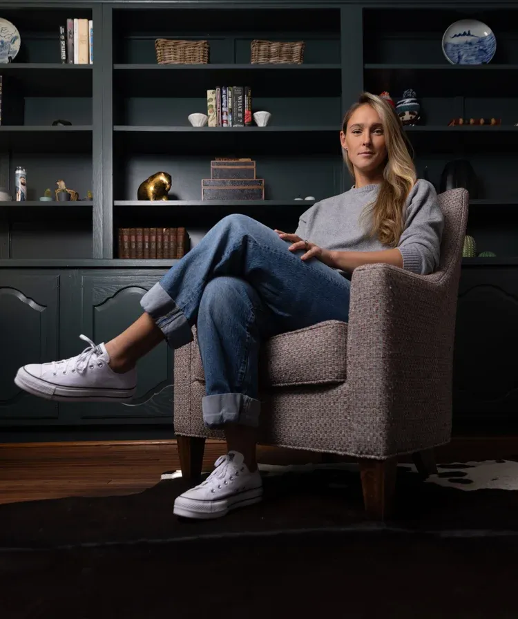 A woman relaxes in a grey armchair wearing casual denim and white sneakers with built-in shelving in the background.