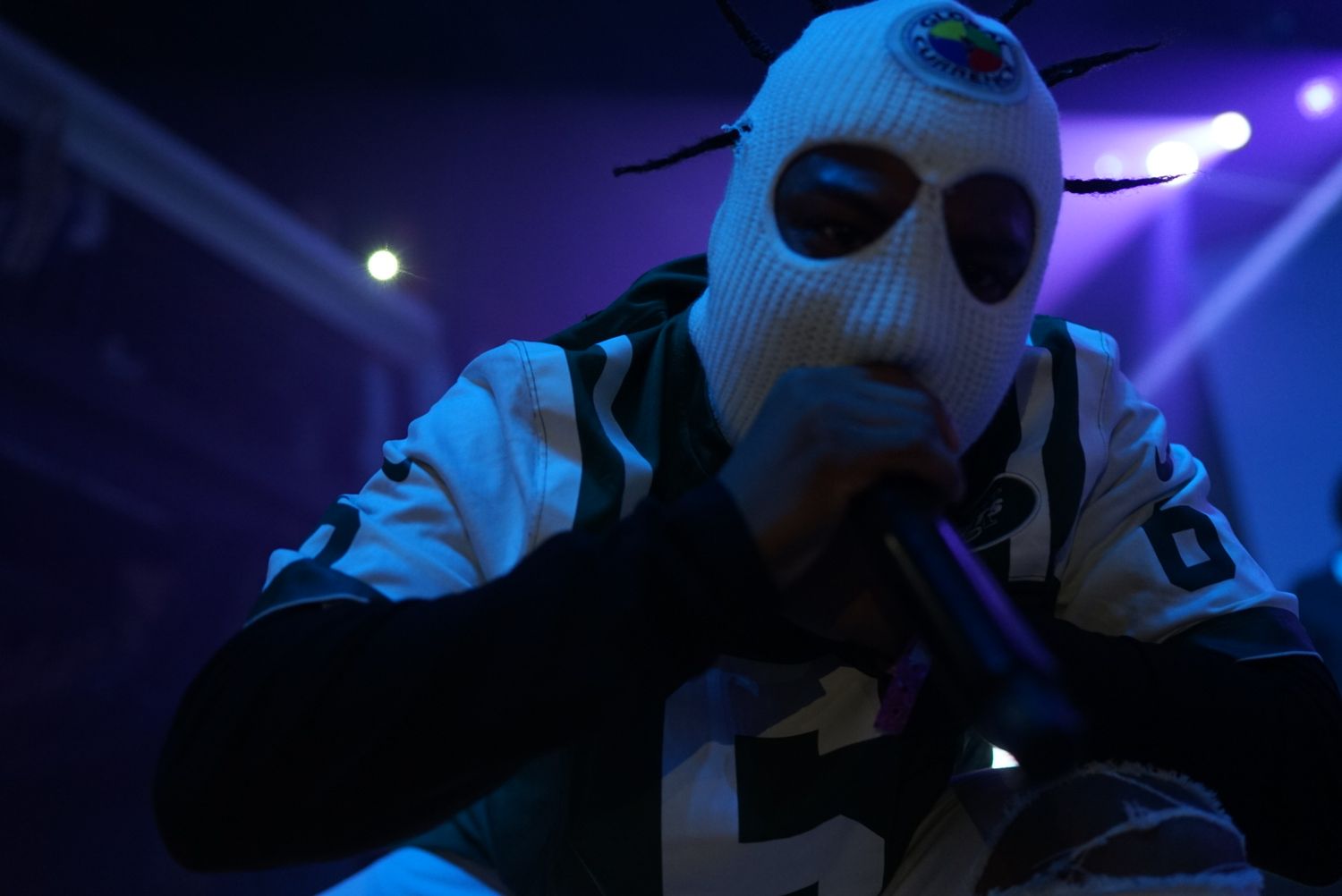 Performer wearing a light blue ski mask performs on stage with purple lighting in the background.