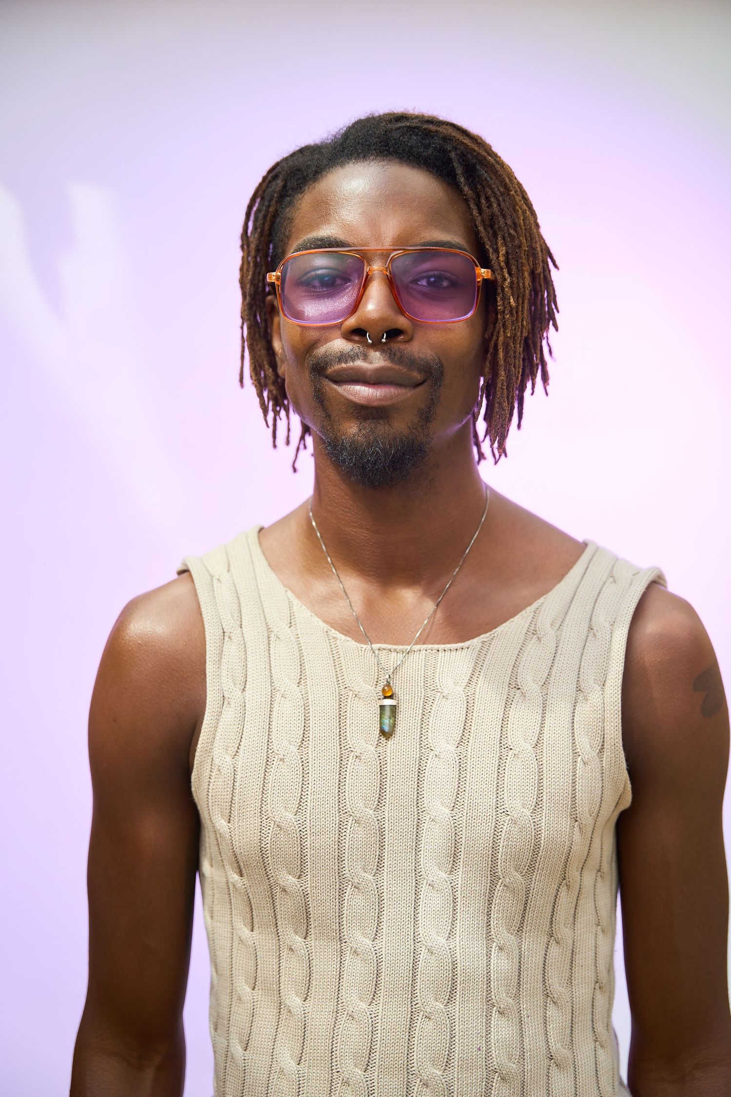 Young Black man with dreadlocks, pink sunglasses, and a crystal necklace wearing a cream knit vest.