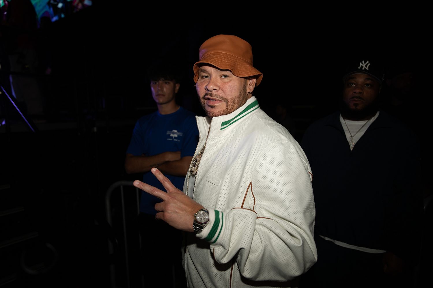 Man in white jacket and orange hat flashing peace sign at a dark indoor event.