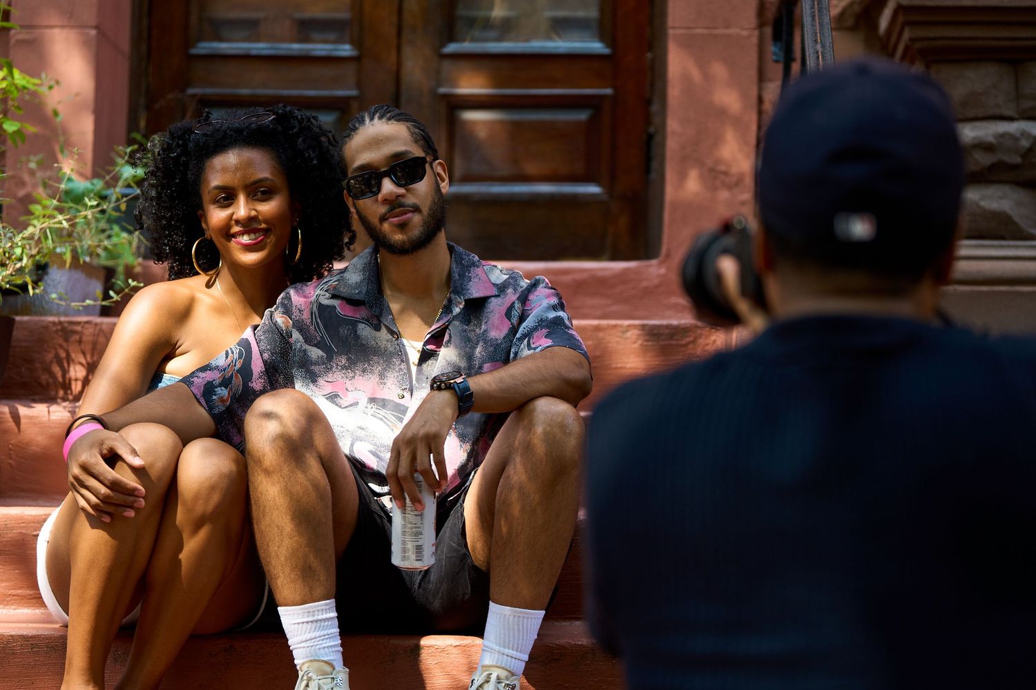 A couple sits together and smiles on a brownstone stoop during a sunny summer day in the city.