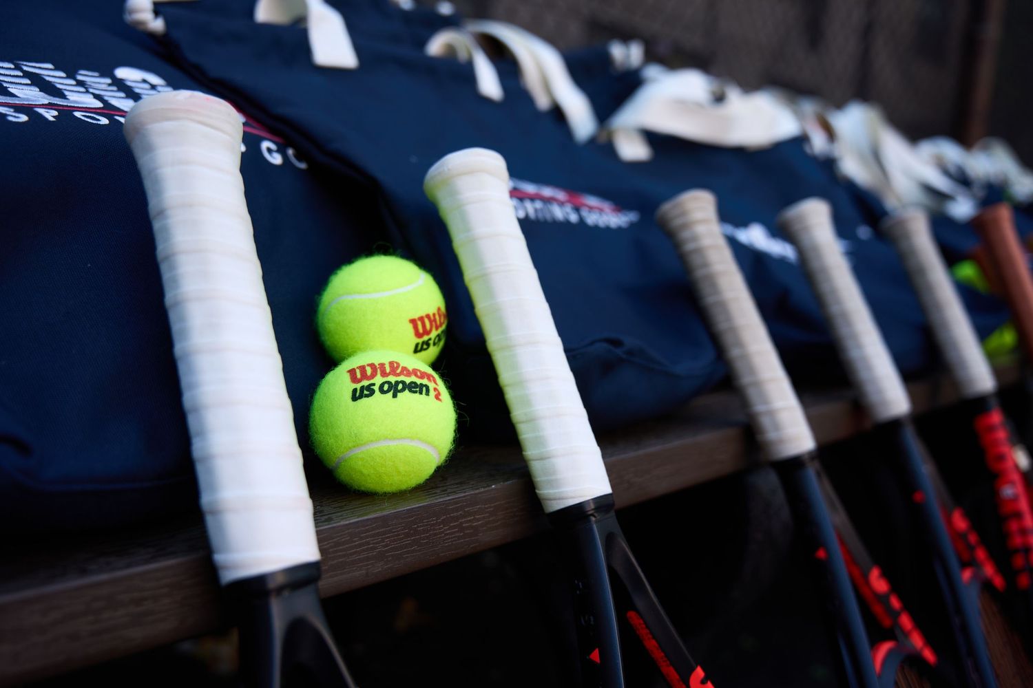 Tennis balls and grip tape rest on a wooden bench alongside tennis rackets at a tennis facility.
