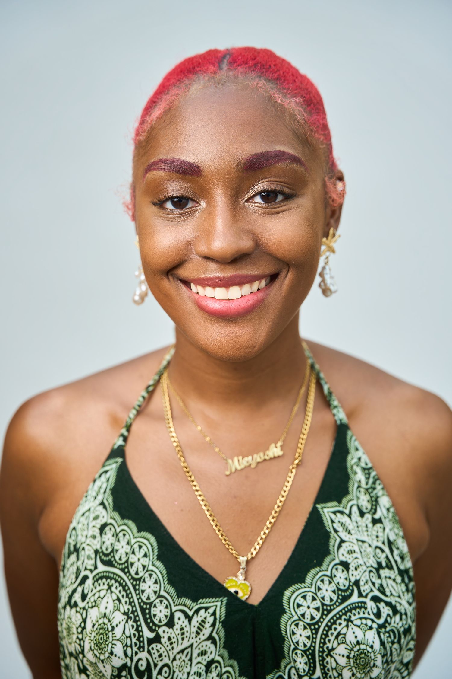 Smiling young woman with pink hair, gold name necklace, and green patterned halter top against gray background.