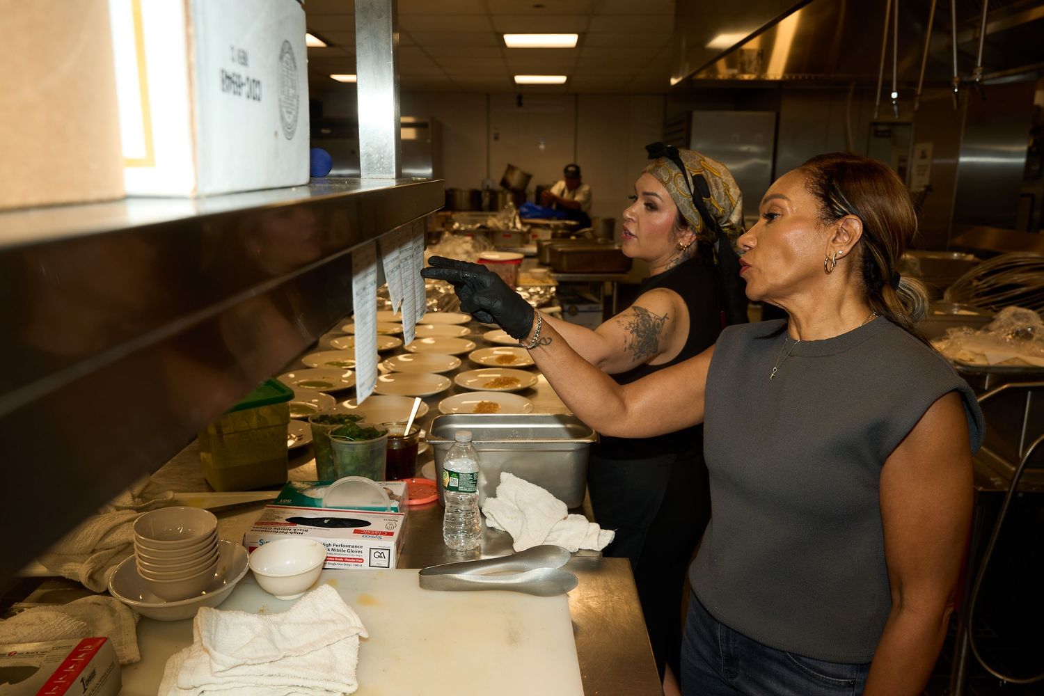 Two women in a busy restaurant kitchen prepare food, one wearing a gray shirt and the other a black apron and green headband.