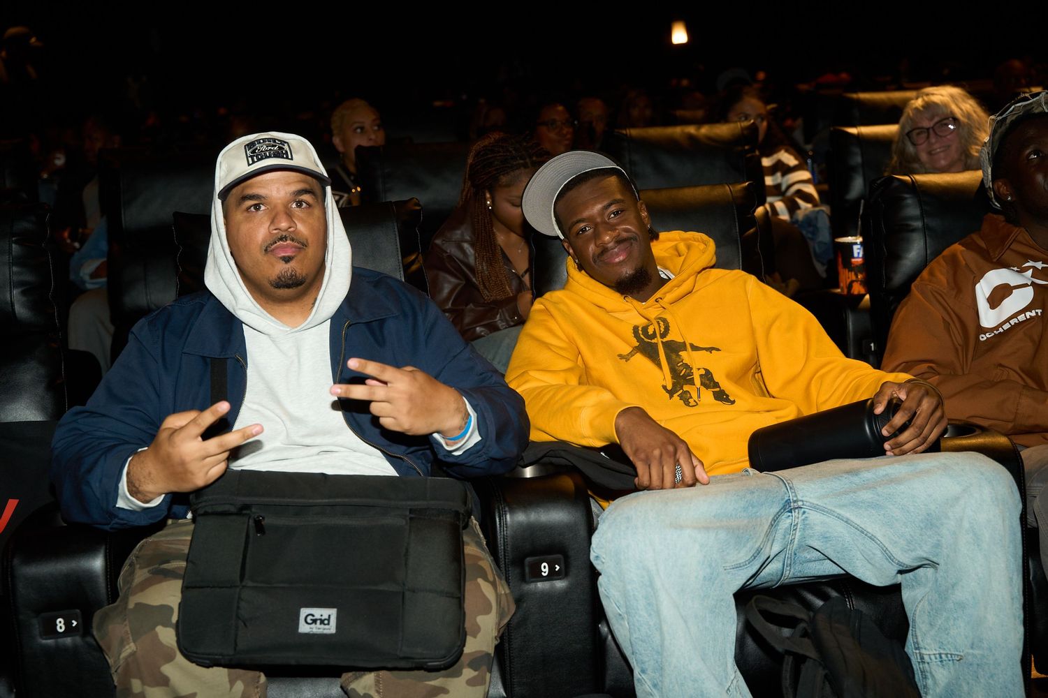 Two men sitting in movie theater seats, one in blue jacket, other in yellow sweatshirt, smiling.
