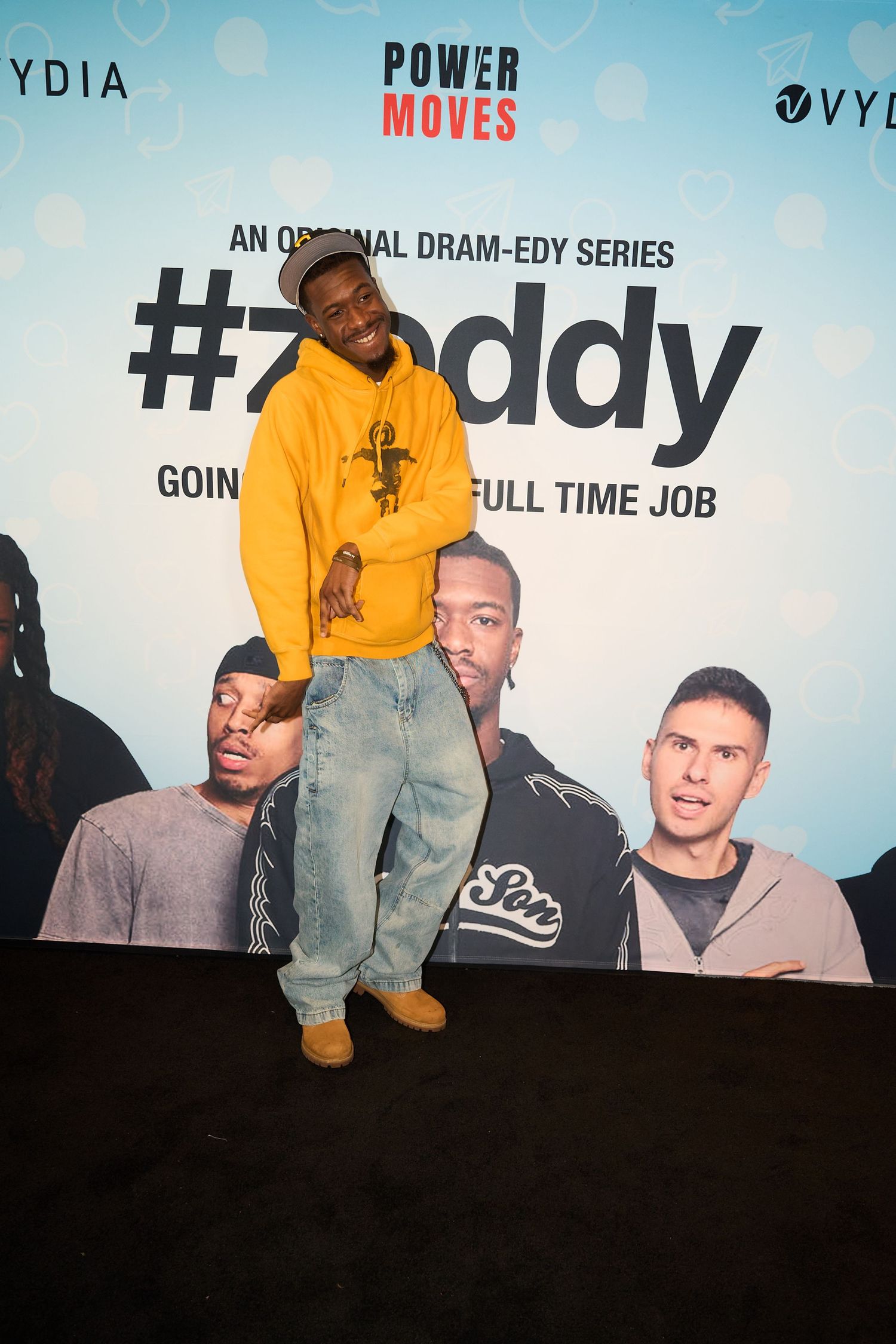 Man in yellow hoodie and baggy jeans poses at #Zeddy dram-edy series premiere event by Power Moves and Vydia.