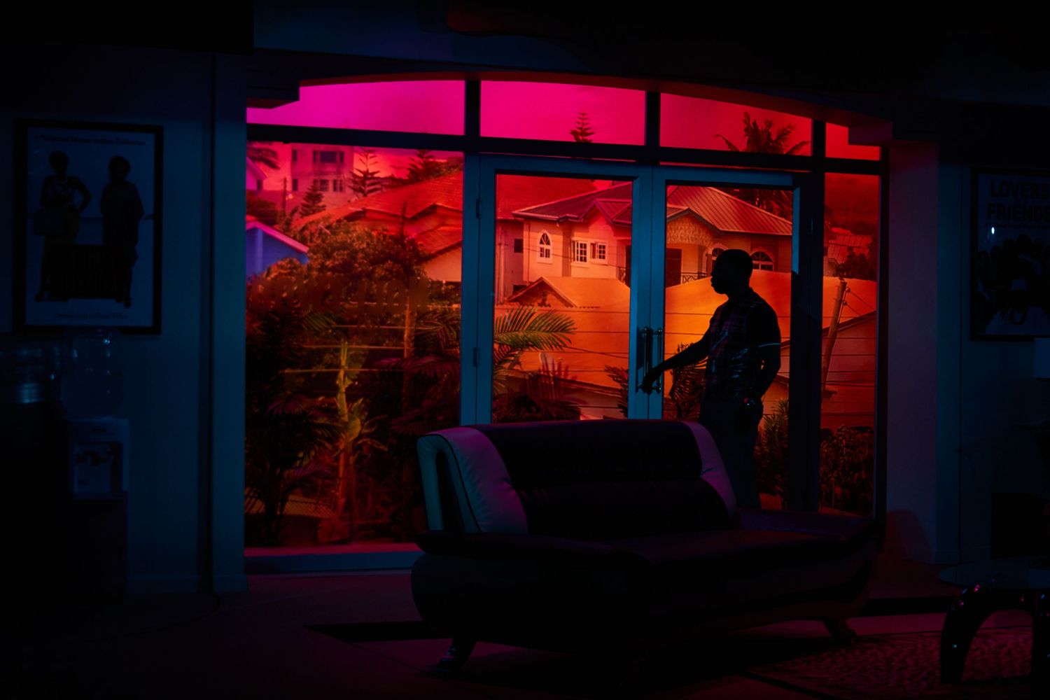 A silhouetted figure stands in front of a window bathed in neon pink light at night.