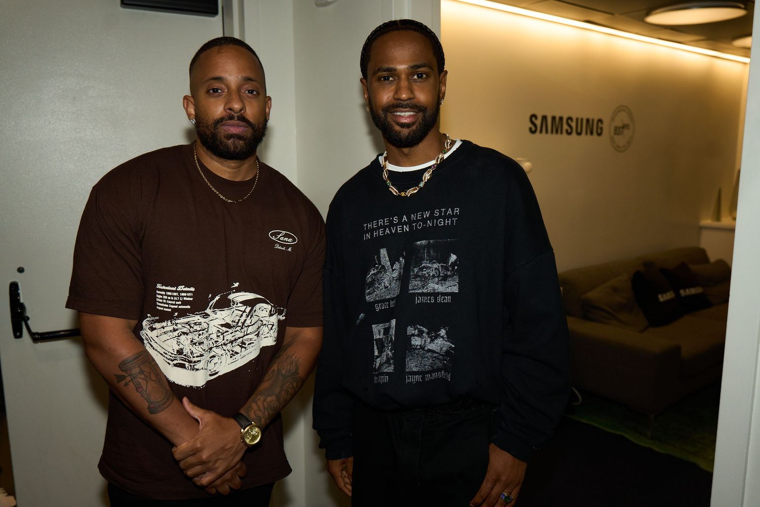 Two men pose together in front of a Samsung logo wearing dark t-shirts in an office setting.