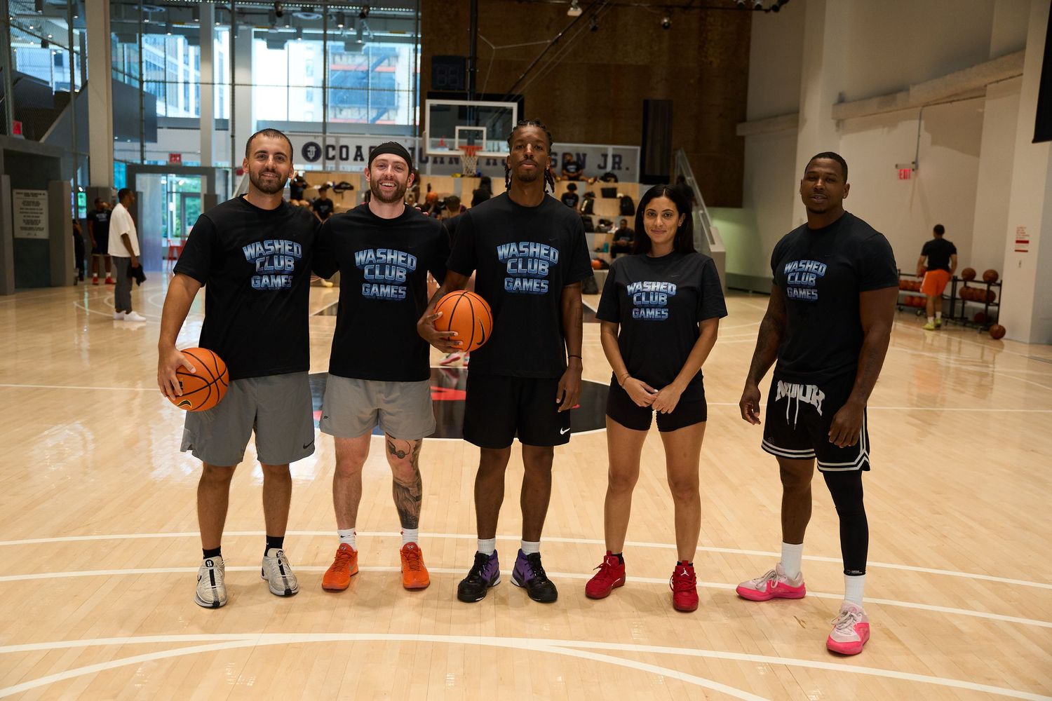Row of players lined up in matching black t-shirts on a basketball court while holding basketballs and posing.