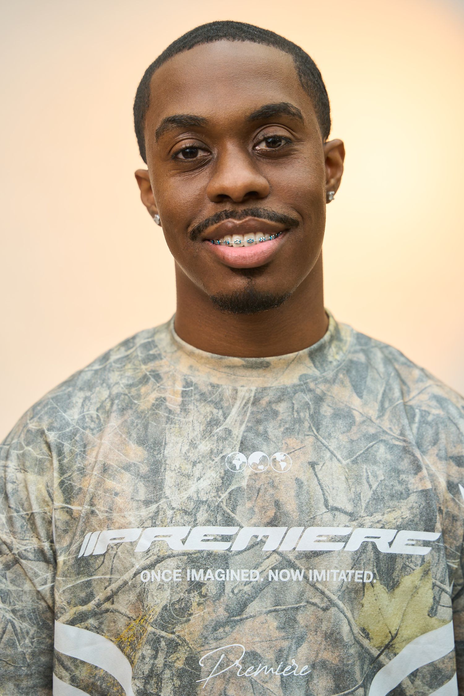 Young man smiling in a camouflage Premiere brand t-shirt against a warm beige background.