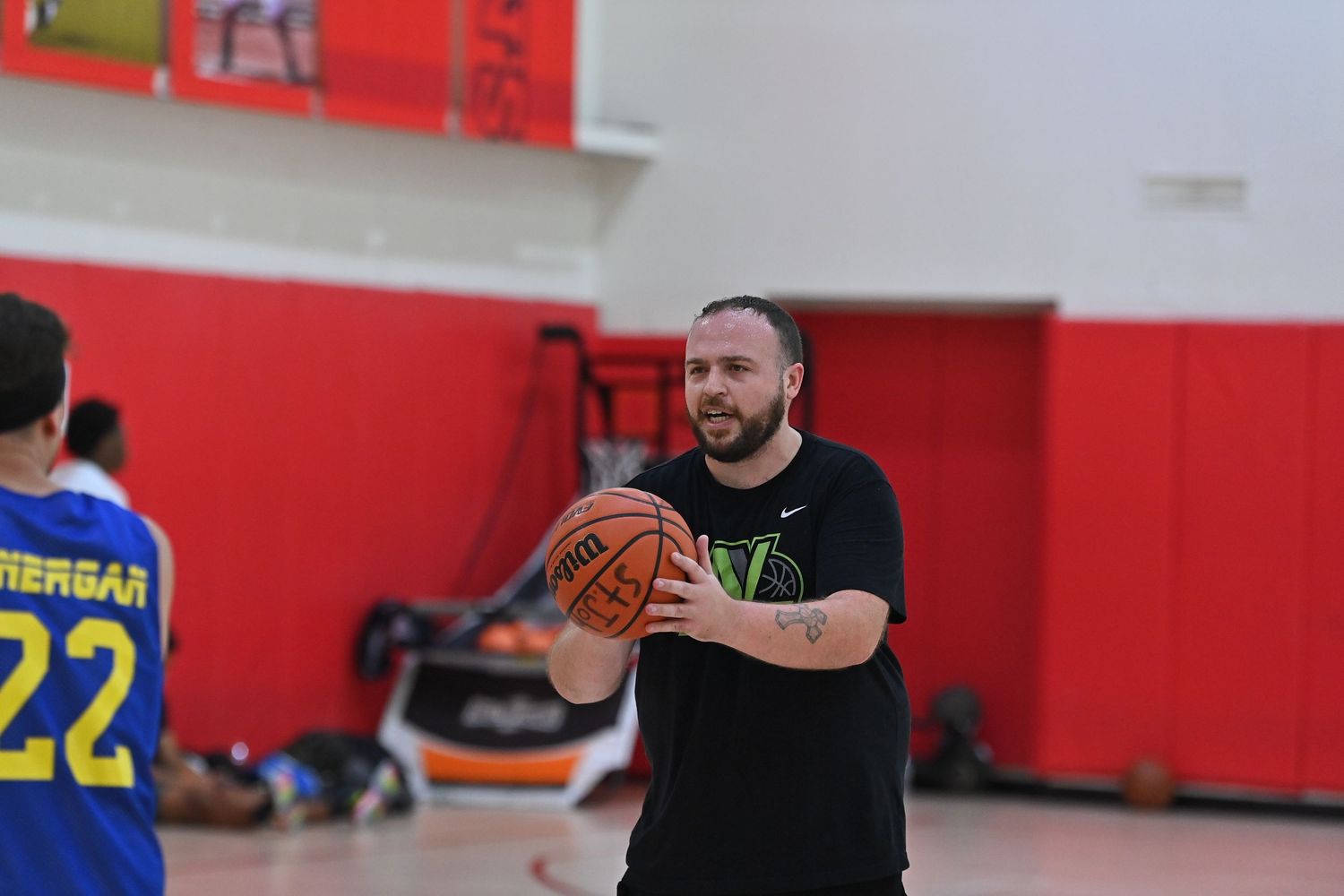 Basketball players compete during an indoor game in a red-walled gymnasium with spectators watching from the sidelines.