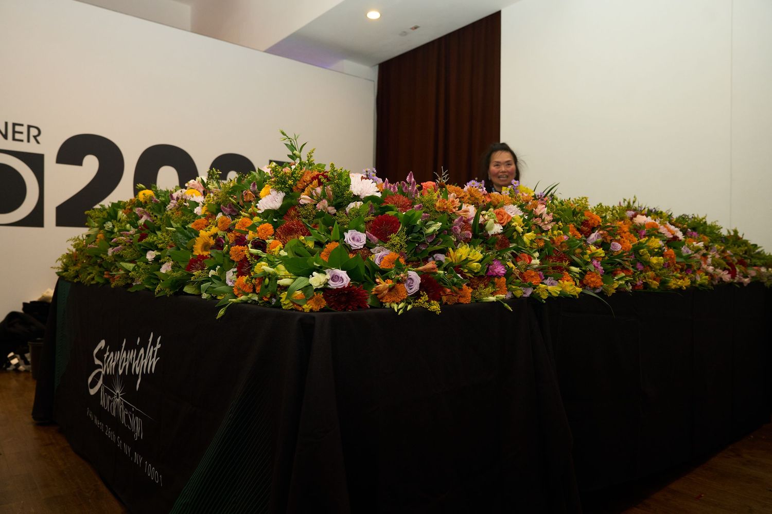 A colorful floral arrangement with orange and yellow flowers adorns a long display table at an event.