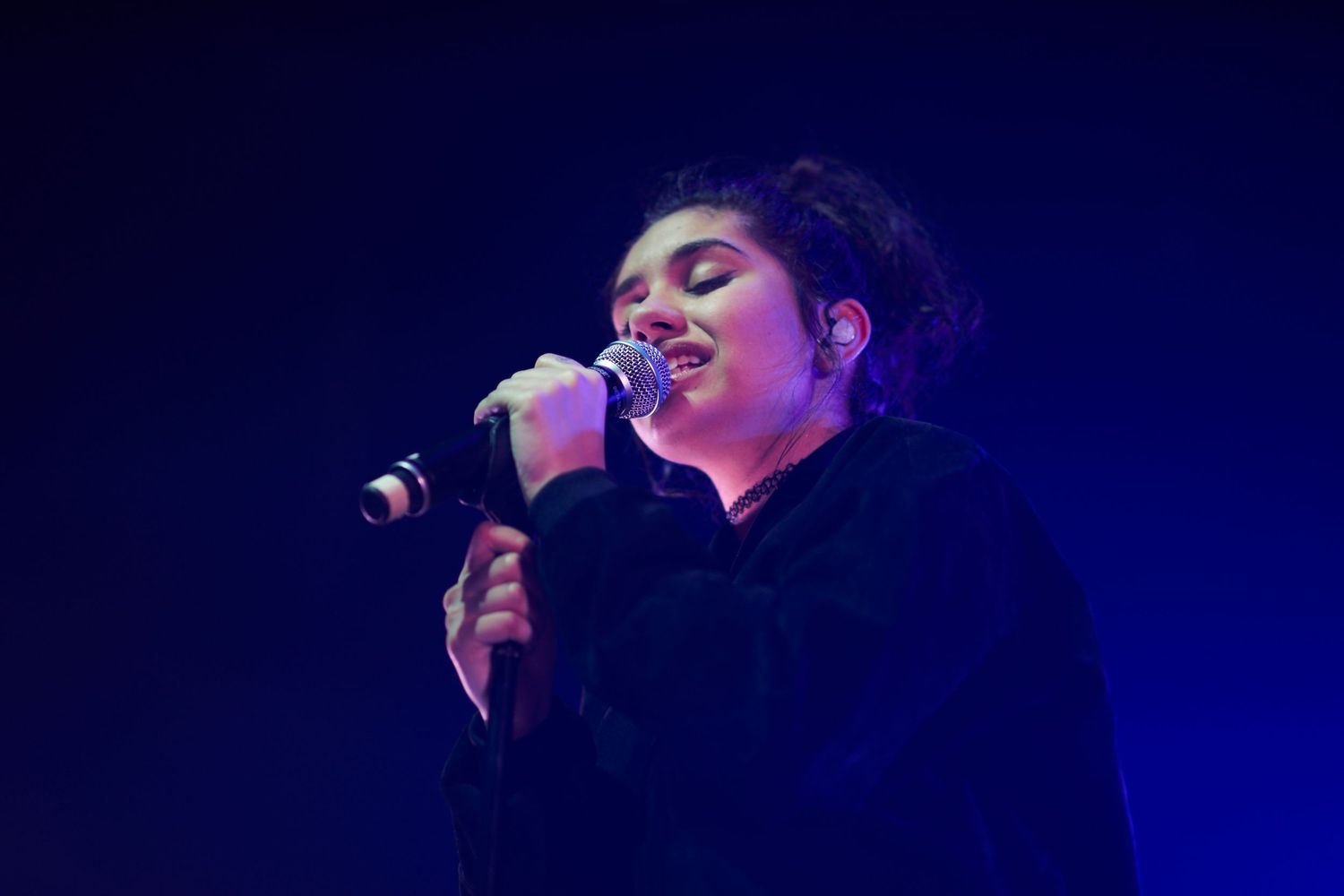 A performer sings passionately into a microphone on a dark stage bathed in blue lighting during a concert performance.