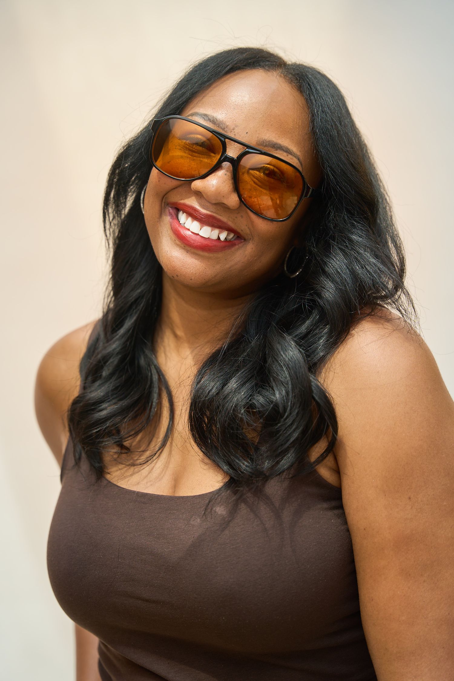 Smiling Black woman with long wavy hair wearing orange tinted aviator sunglasses and a brown tank top.