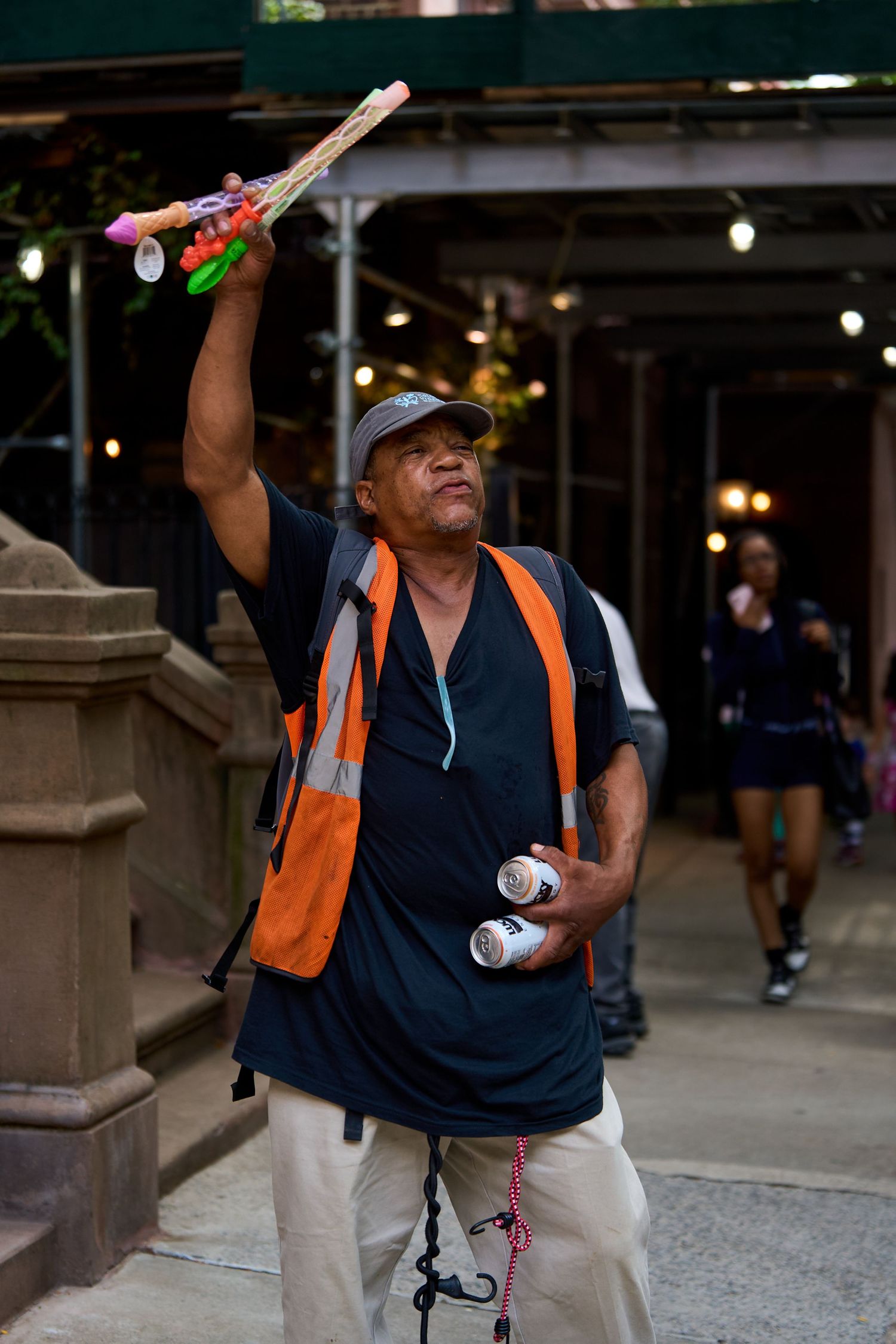 A street vendor in an orange safety vest holds up novelty toys while working outdoors.