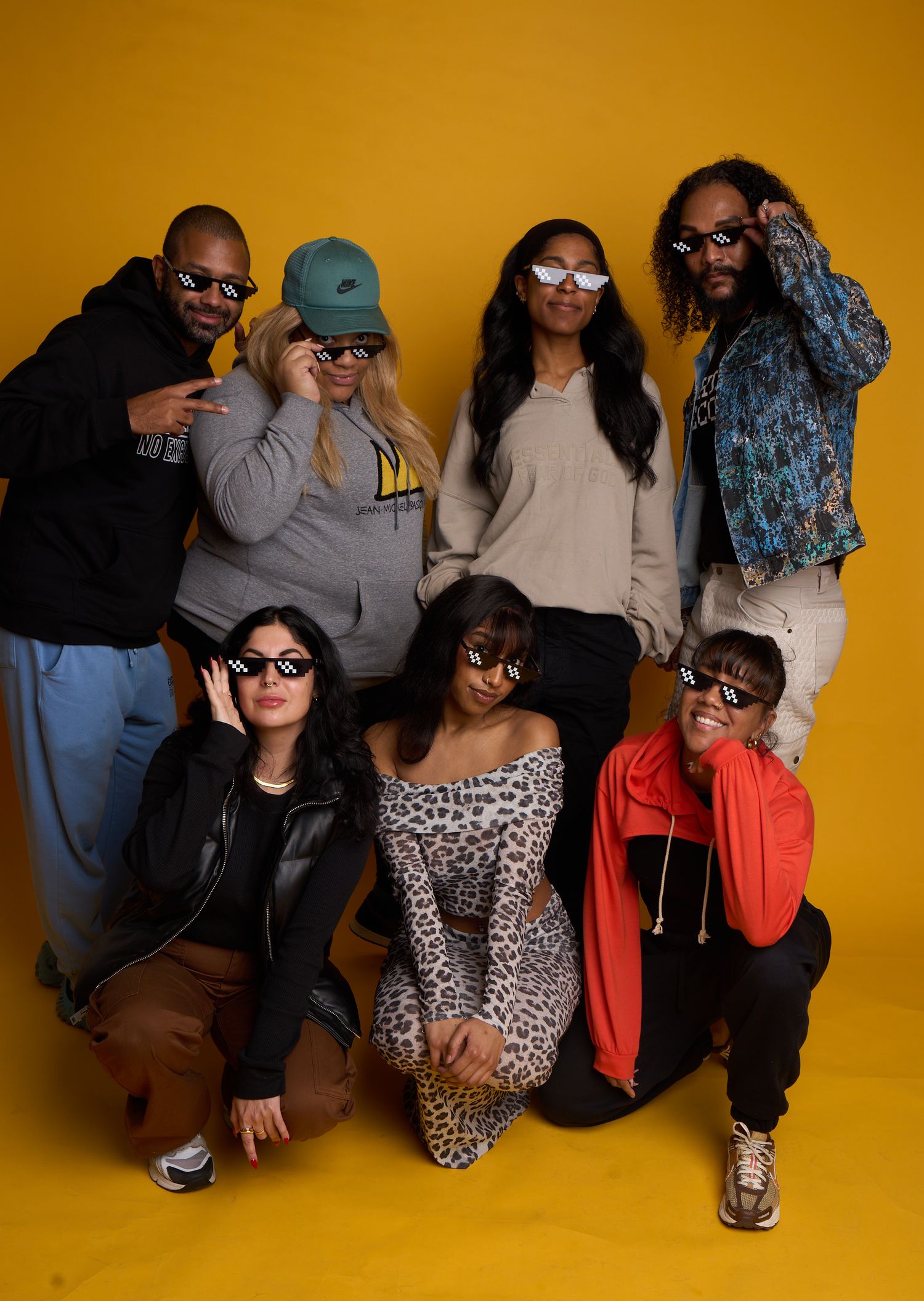 Group of seven friends striking stylish poses in casual outfits against a vibrant yellow background in a studio setting.