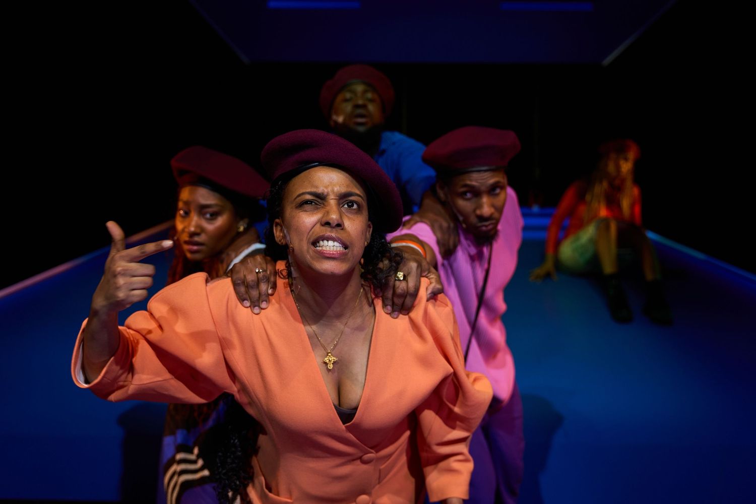 Performers in vibrant orange and red outfits dance energetically on a stage with blue lighting during a theatrical show.