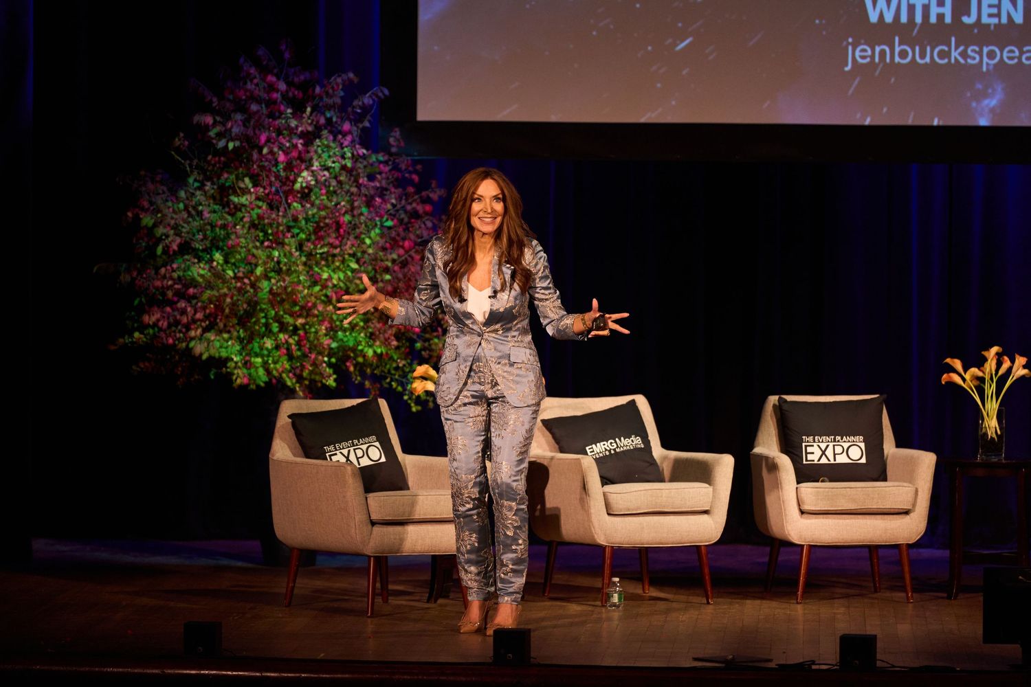 Speakers engage in conversation on stage with white chairs and dark backdrop at professional conference event.