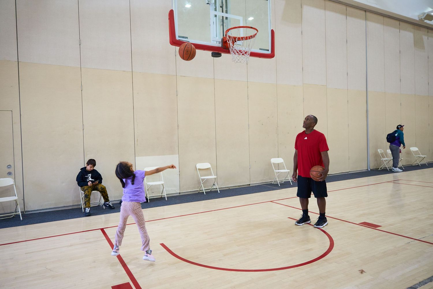 A person in red plays basketball in a gymnasium with white walls and a basketball hoop visible.