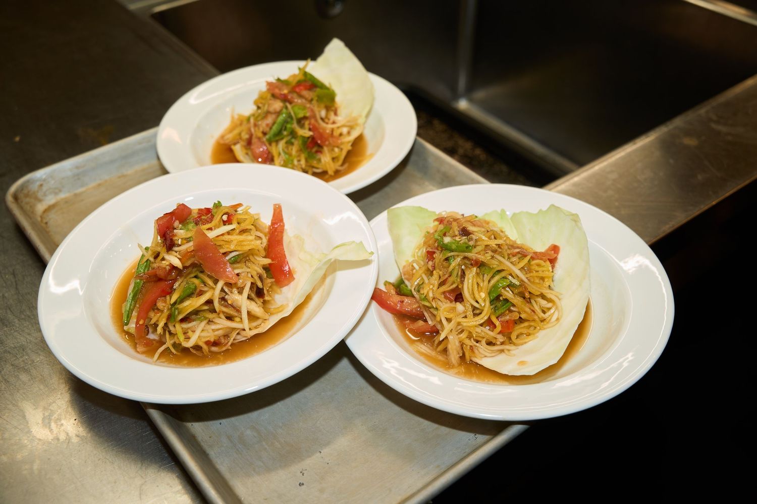 Three white plates of fresh papaya salad with shredded vegetables, tomatoes, and herbs served on cabbage leaves atop a metal serving tray.