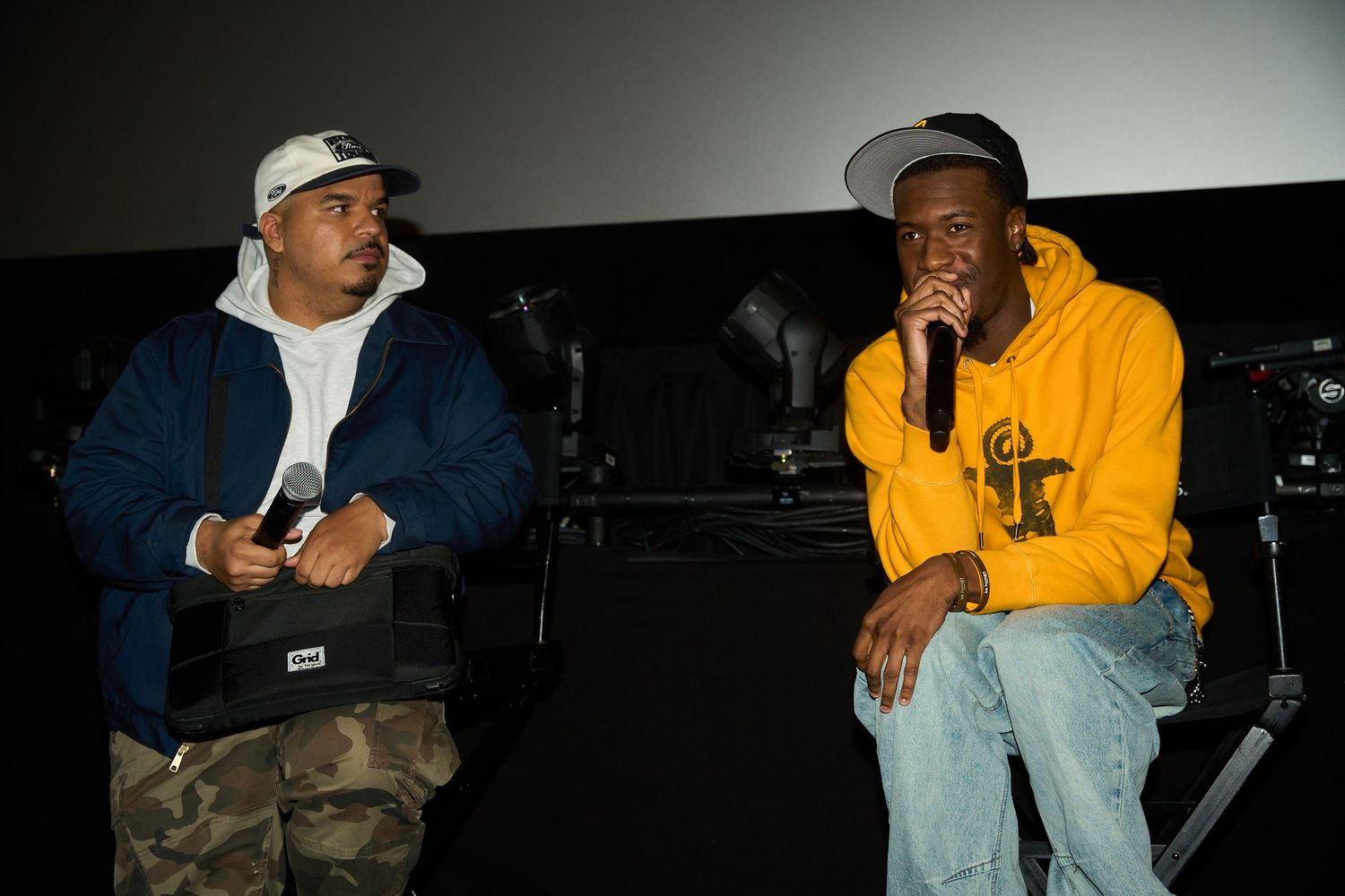 Two men sit on stage at an indoor event, one in a navy jacket with a mic, the other in a yellow hoodie speaking.