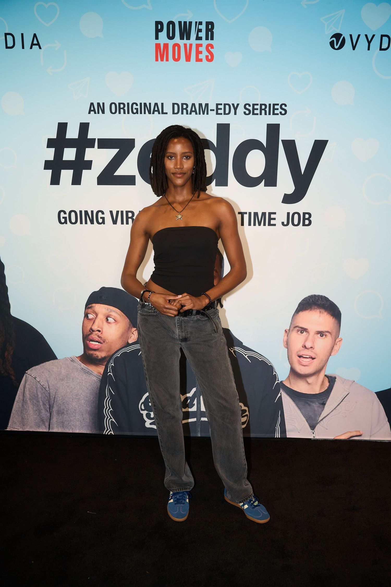 A young woman poses at the #Zeddy dram-edy series premiere event backdrop wearing a black tube top and gray jeans.
