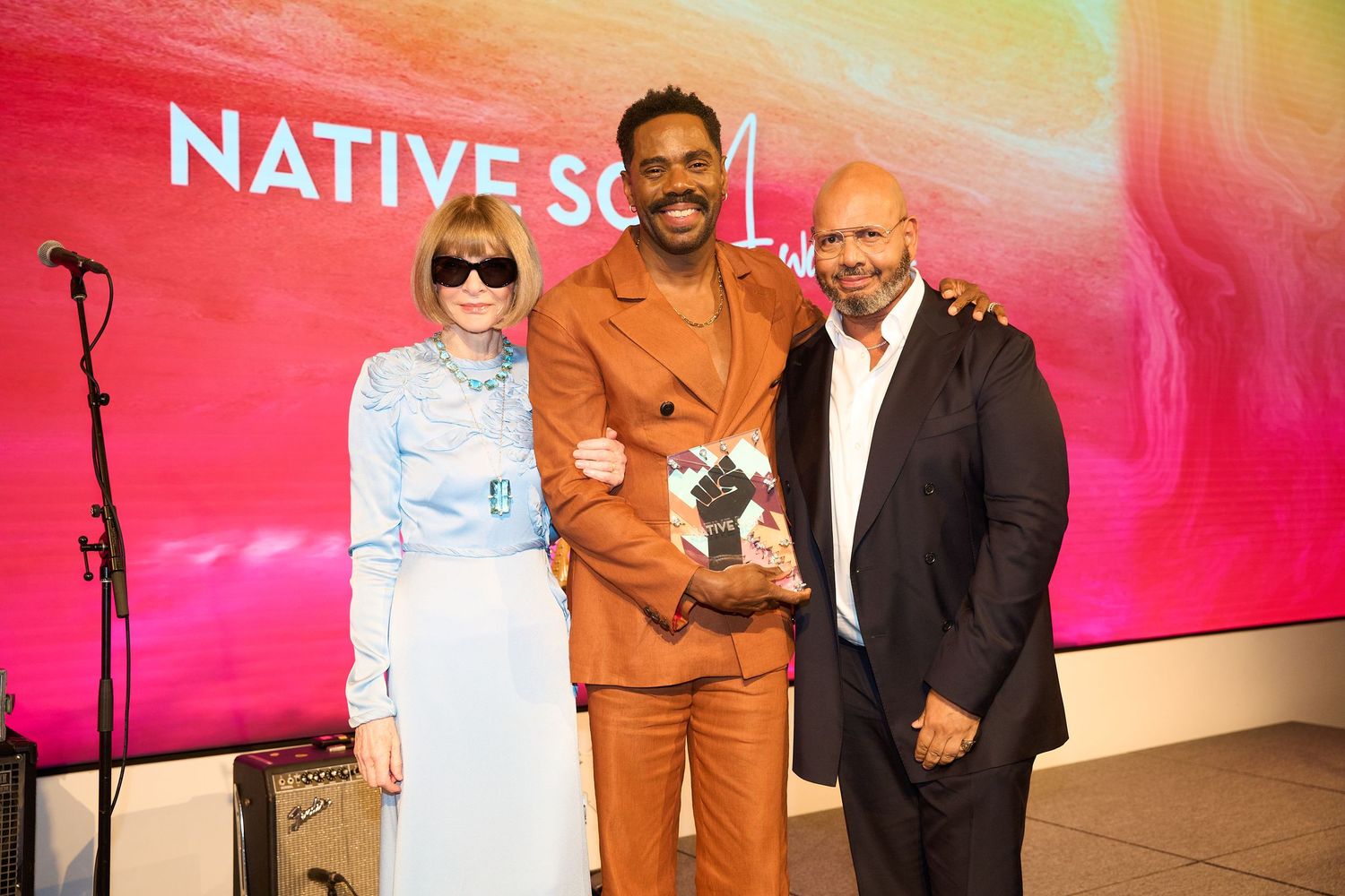 Three people (Anna Wintor, Colman Domingo, Emil) stand together smiling against a vibrant pink and orange gradient backdrop at an event.