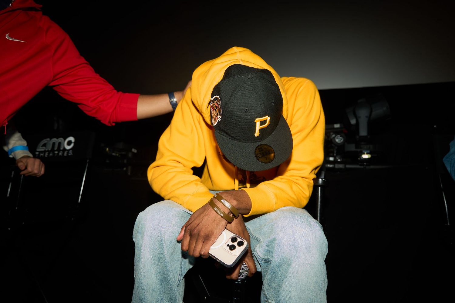 Man in yellow hoodie and black Pittsburgh Pirates cap sits with head down holding a phone at AMC theater.