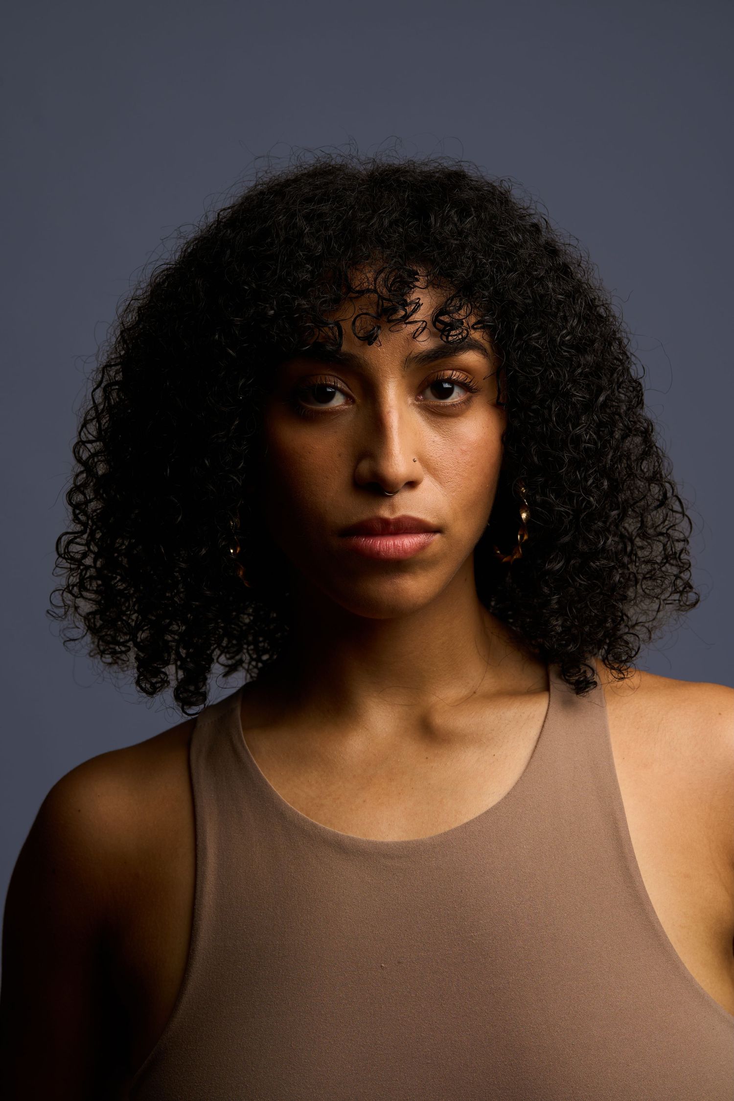 Young woman with curly black hair wearing a beige tank top, posing against a gray background.