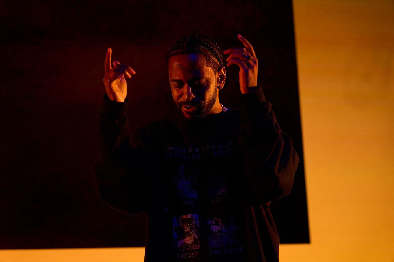 A person gestures with raised hands in a dramatic silhouette against an orange-lit backdrop.