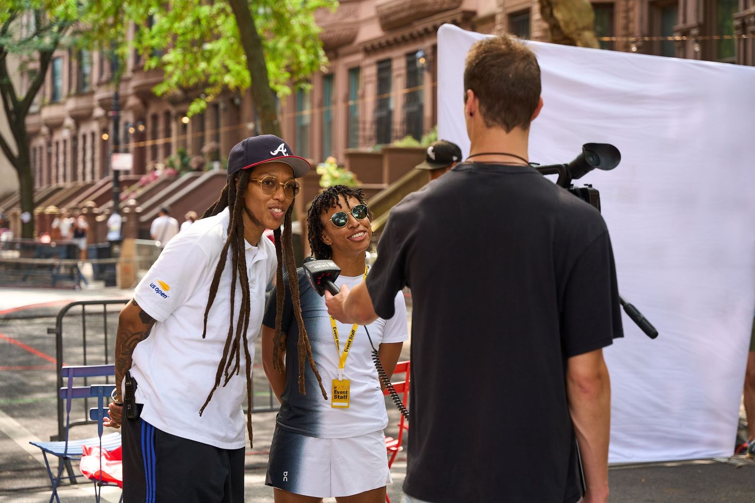 People having a conversation on an urban sidewalk in front of a white backdrop during a sunny day.