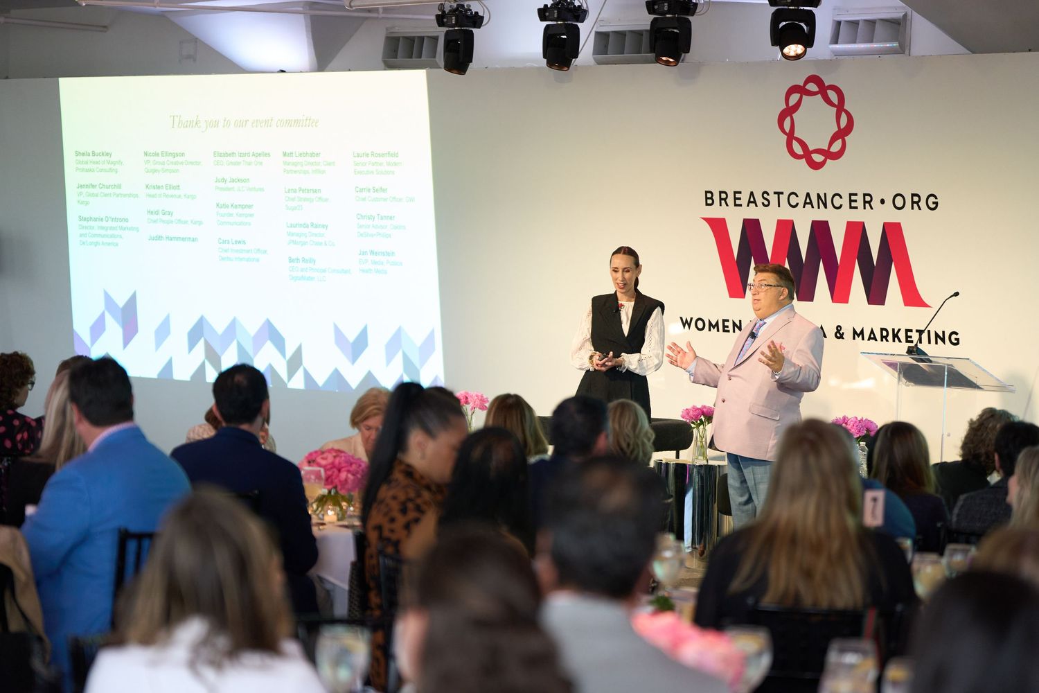 Conference presentation in a modern meeting room with speakers on stage and audience seated in rows.