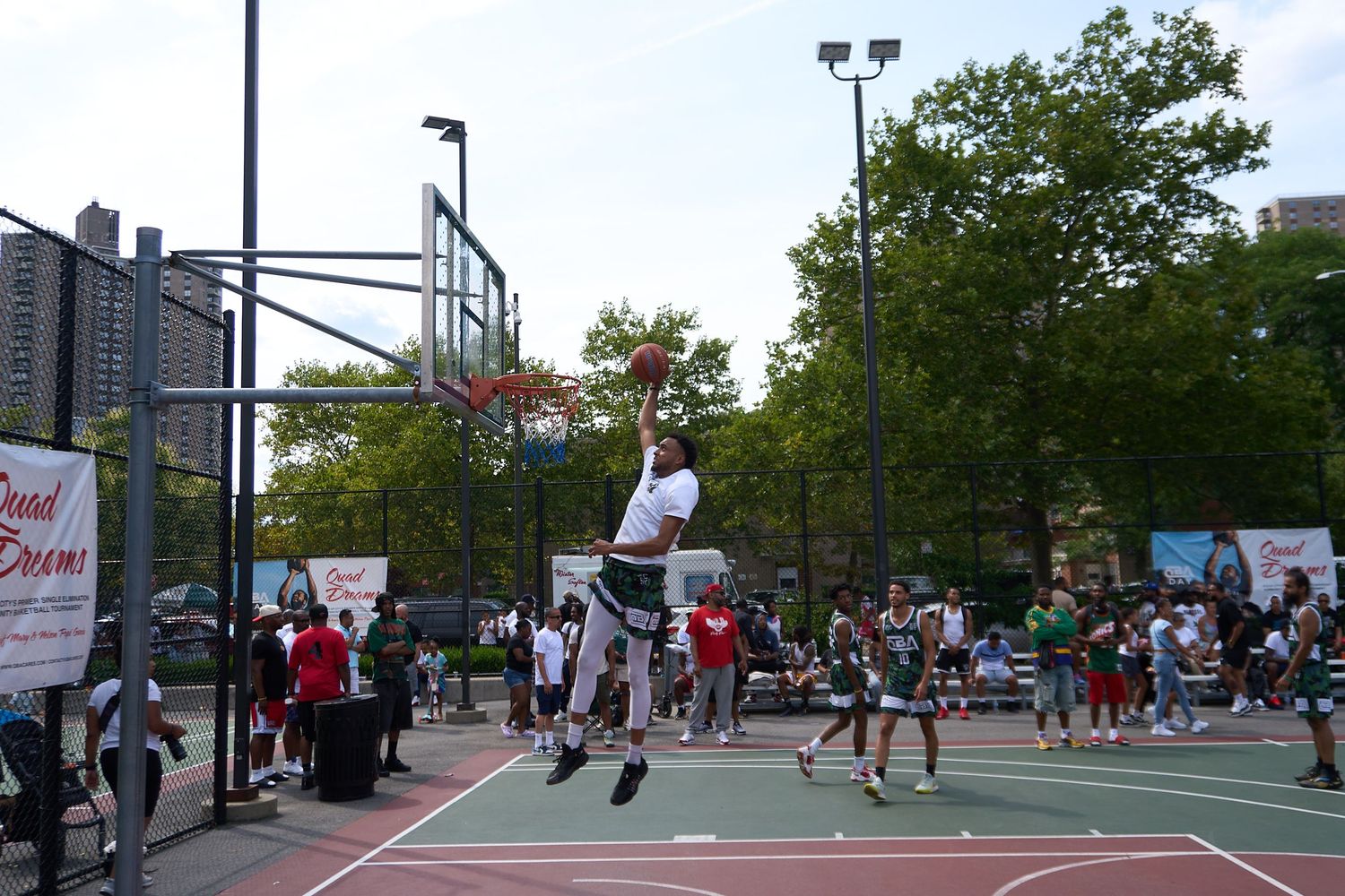 People play basketball on an outdoor court with spectators watching from the sidelines during a daytime game.