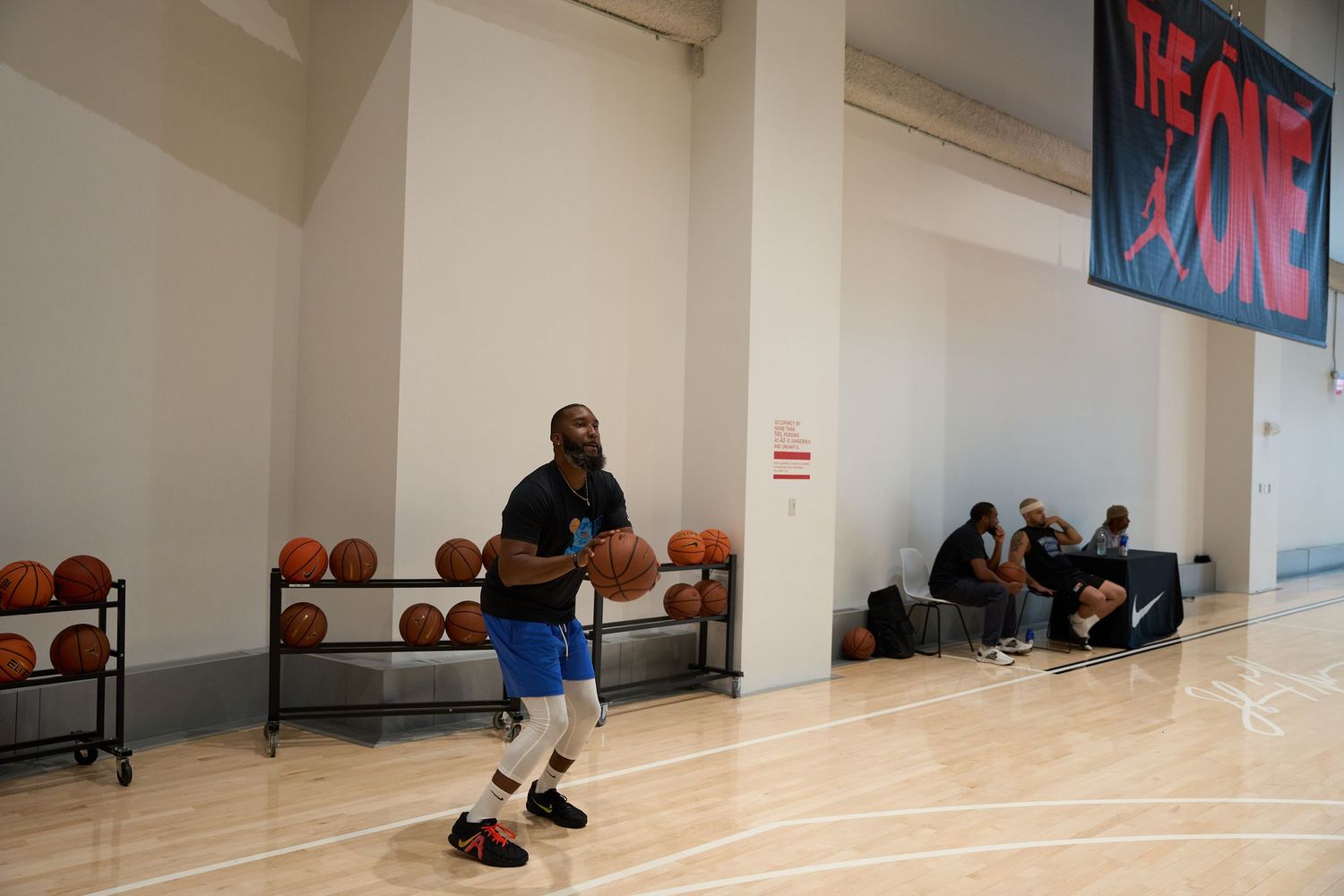 Basketball players practice shooting and drills on a polished wooden court in a bright indoor gymnasium.
