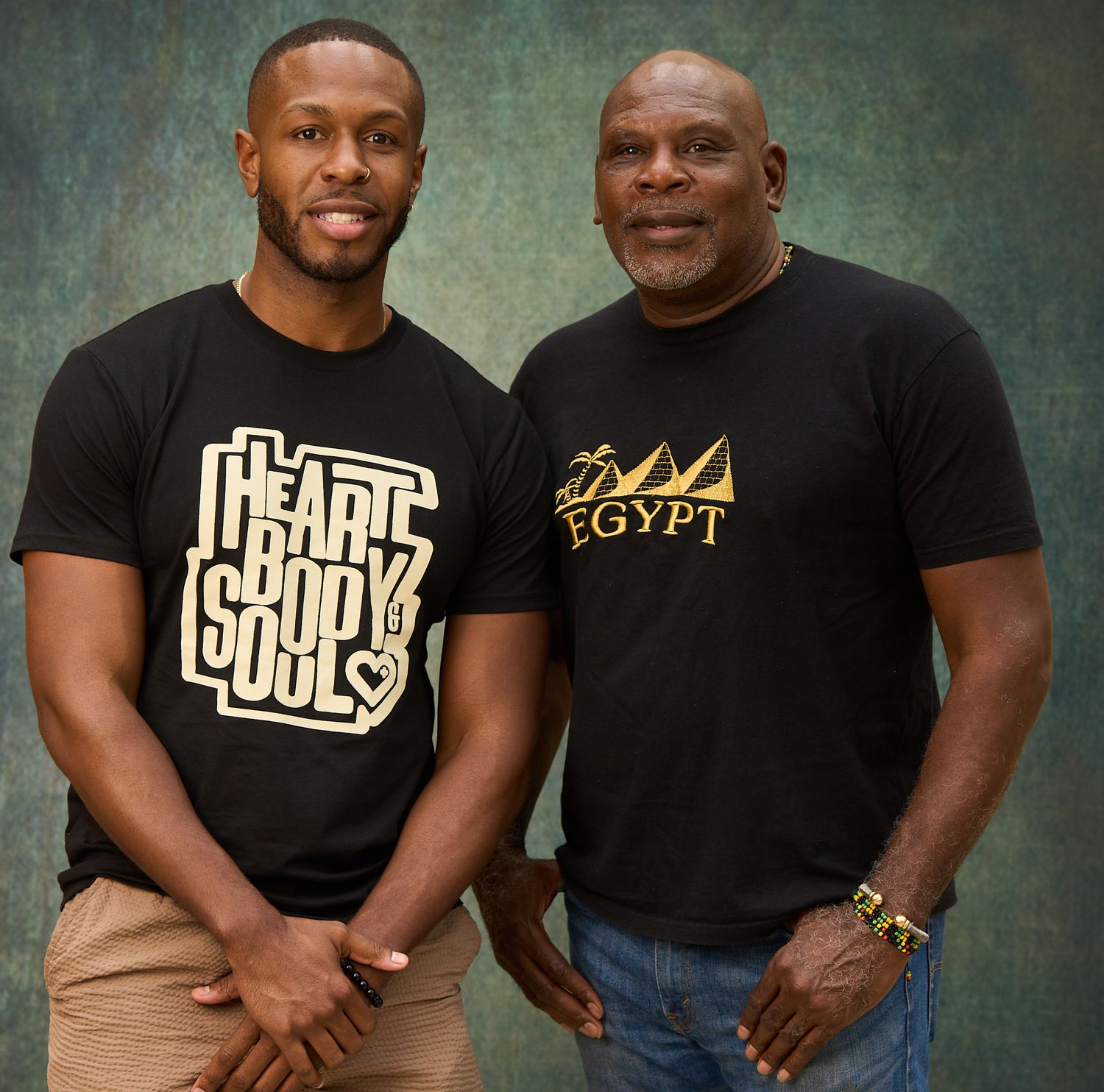 Two men in black t-shirts with graphic designs pose together for a studio portrait against a textured background.