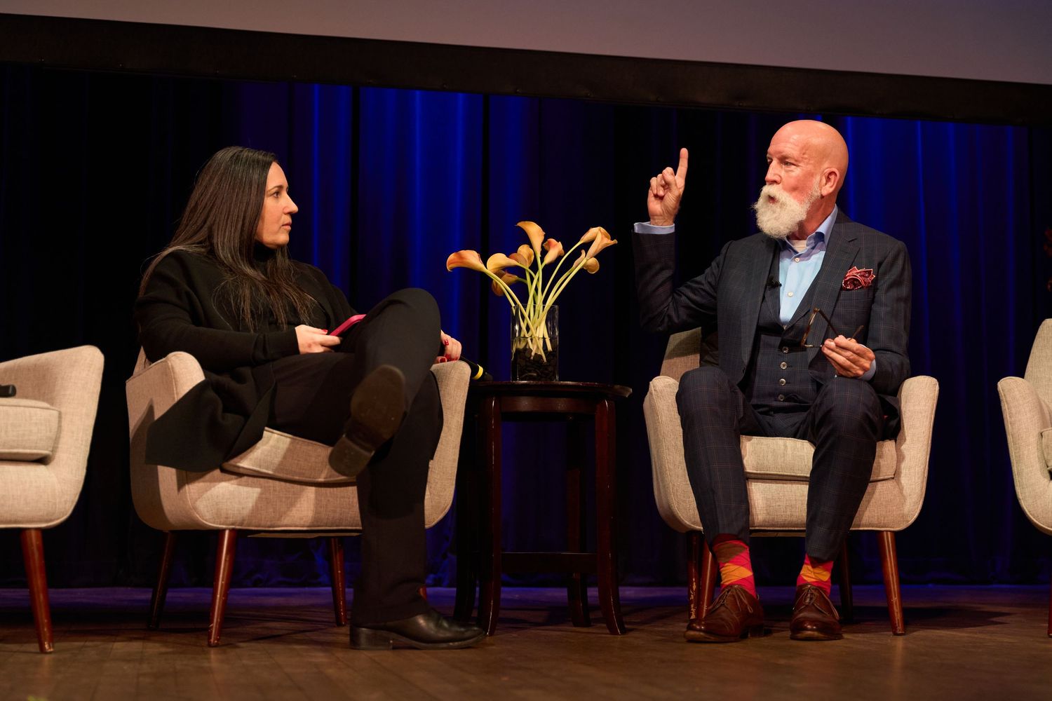 Conference speakers engaged in conversation while seated in comfortable armchairs on a dimly lit stage.