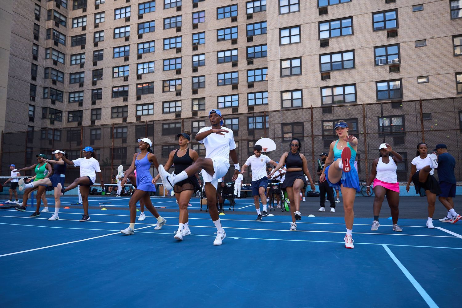 People practicing tennis drills on multiple blue courts between city apartment buildings.