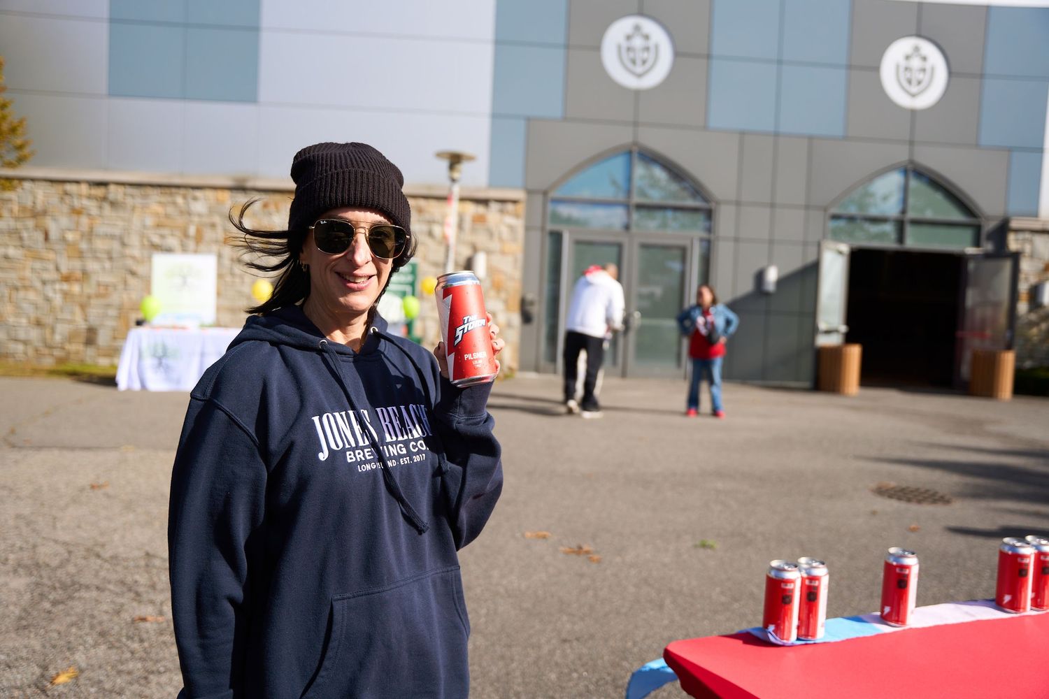 A person in sunglasses and dark clothing stands next to a red table with drinks outdoors.
